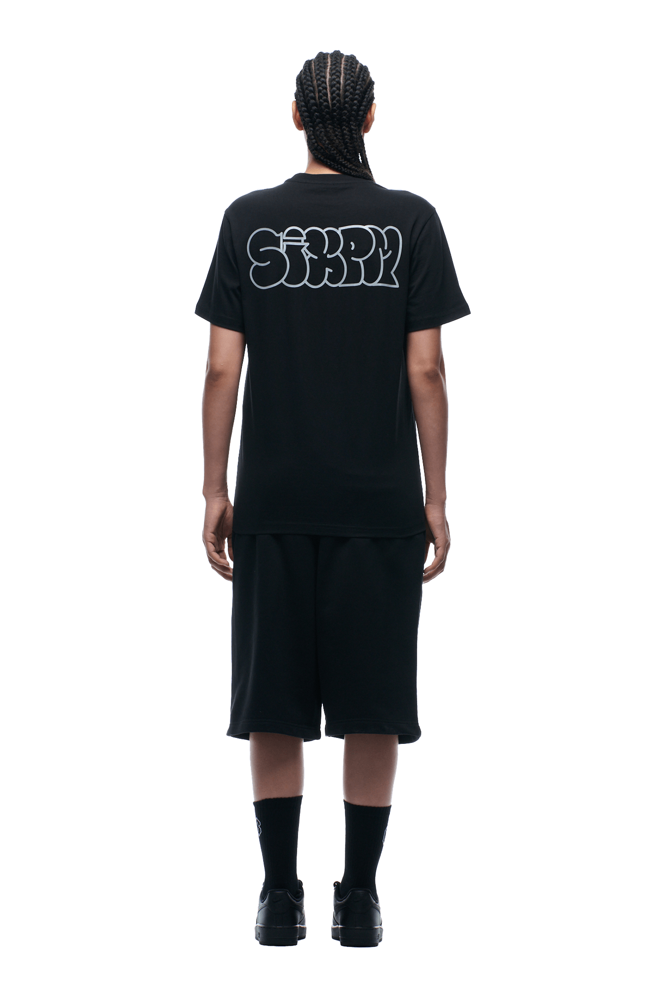 A person faces away from the camera wearing the 6PM REFLECTIVE LOGO TEE BLACK, paired with black shorts, socks, and shoes, all set against a plain background.