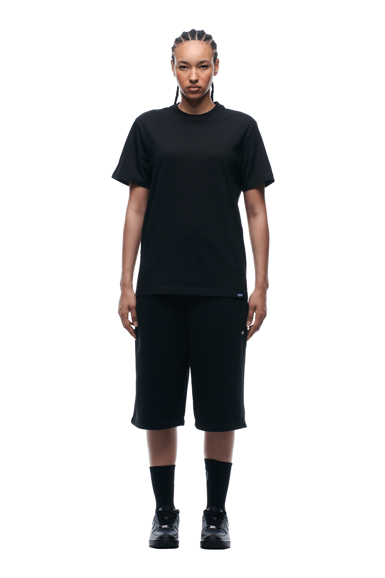 A person stands facing forward, wearing a 6PM REFLECTIVE LOGO TEE BLACK, loose black knee-length shorts, black socks, and sneakers with neat cornrows. Background is transparent.