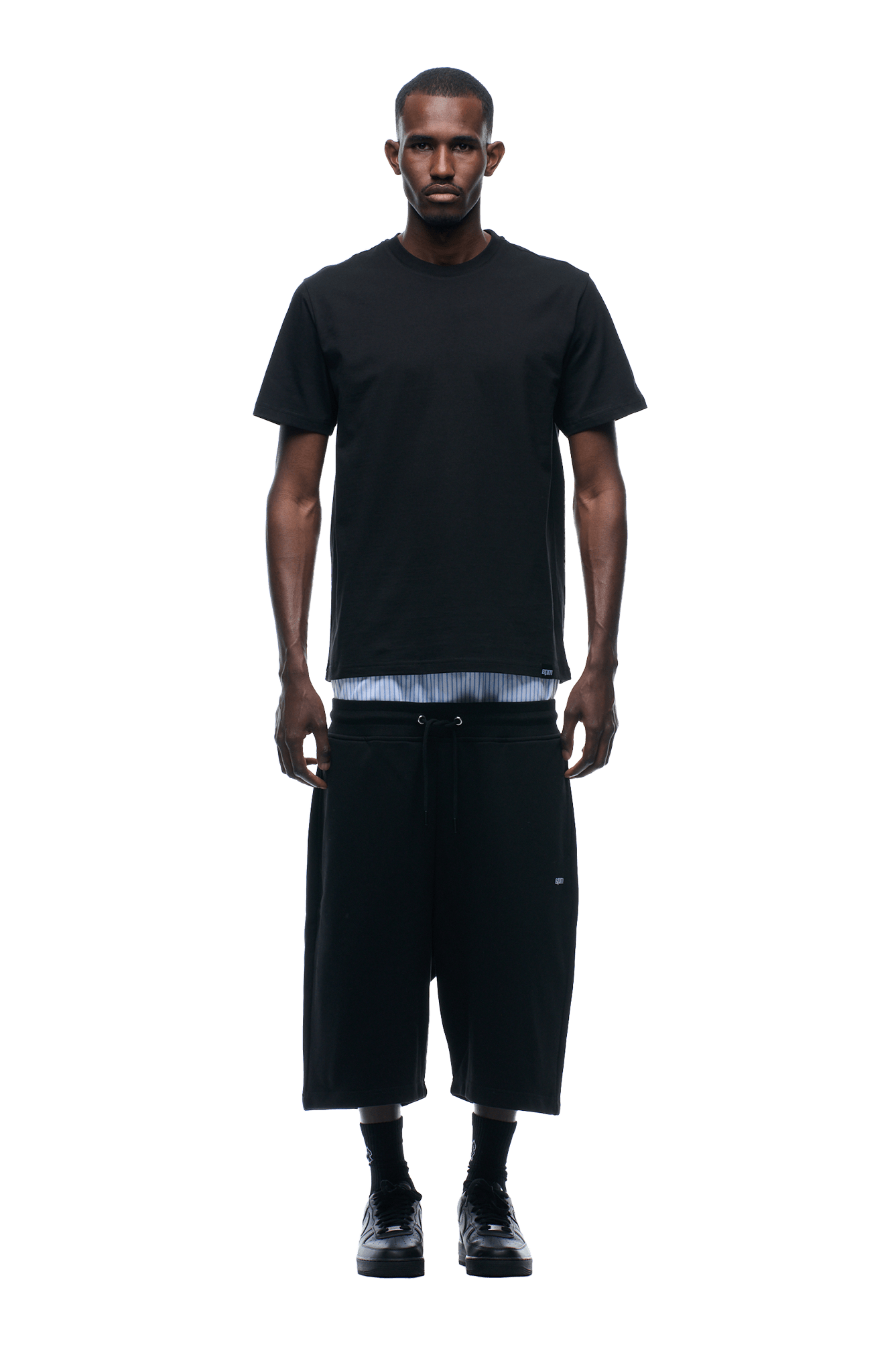 A man faces forward against a plain green background, wearing the 6PM REFLECTIVE LOGO TEE BLACK over a striped undershirt, with wide black shorts, black socks, and black shoes.