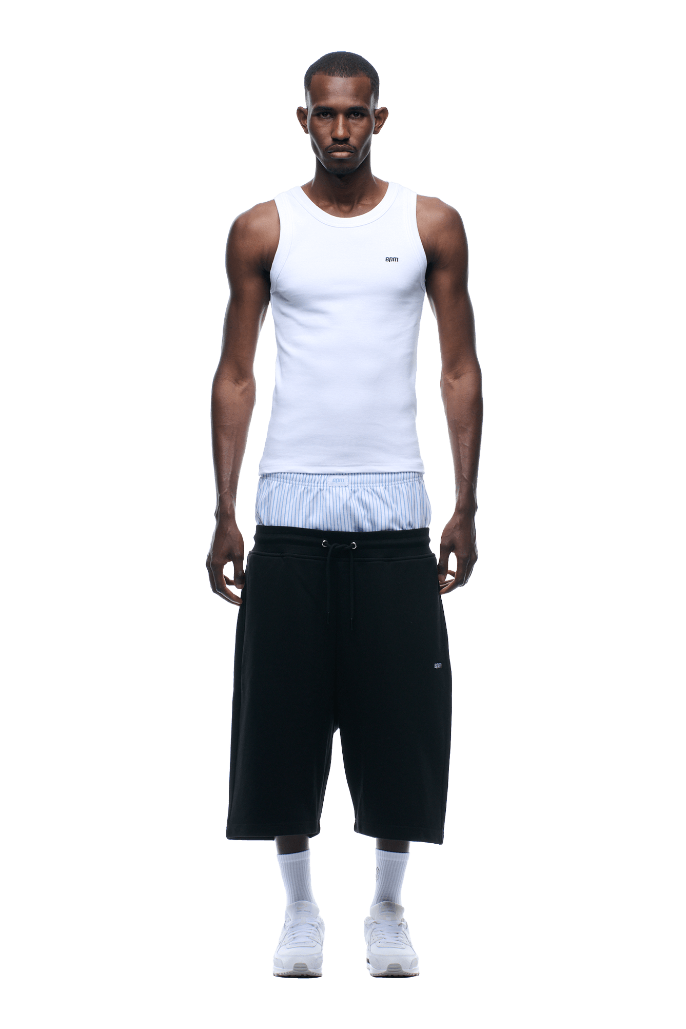A man stands facing forward against a plain green background, wearing 6PM BOXERS SHORTS BLACK, a white tank top, blue and white striped shirt underneath, white socks, and white sneakers.