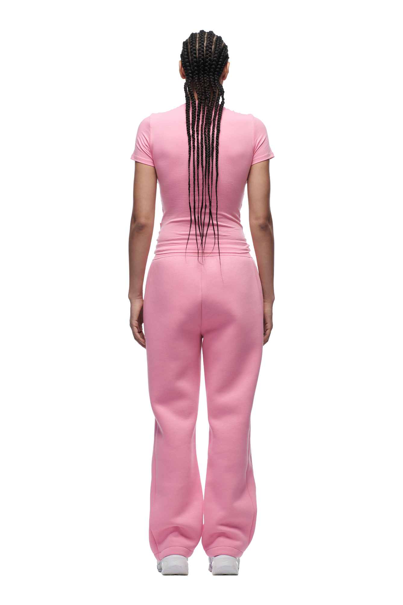 A person with long, braided hair stands facing away in a fitted 6PM WMNS T-SHIRT BUBBLEGLOW, loose pink sweatpants, and white sneakers against a plain white background.