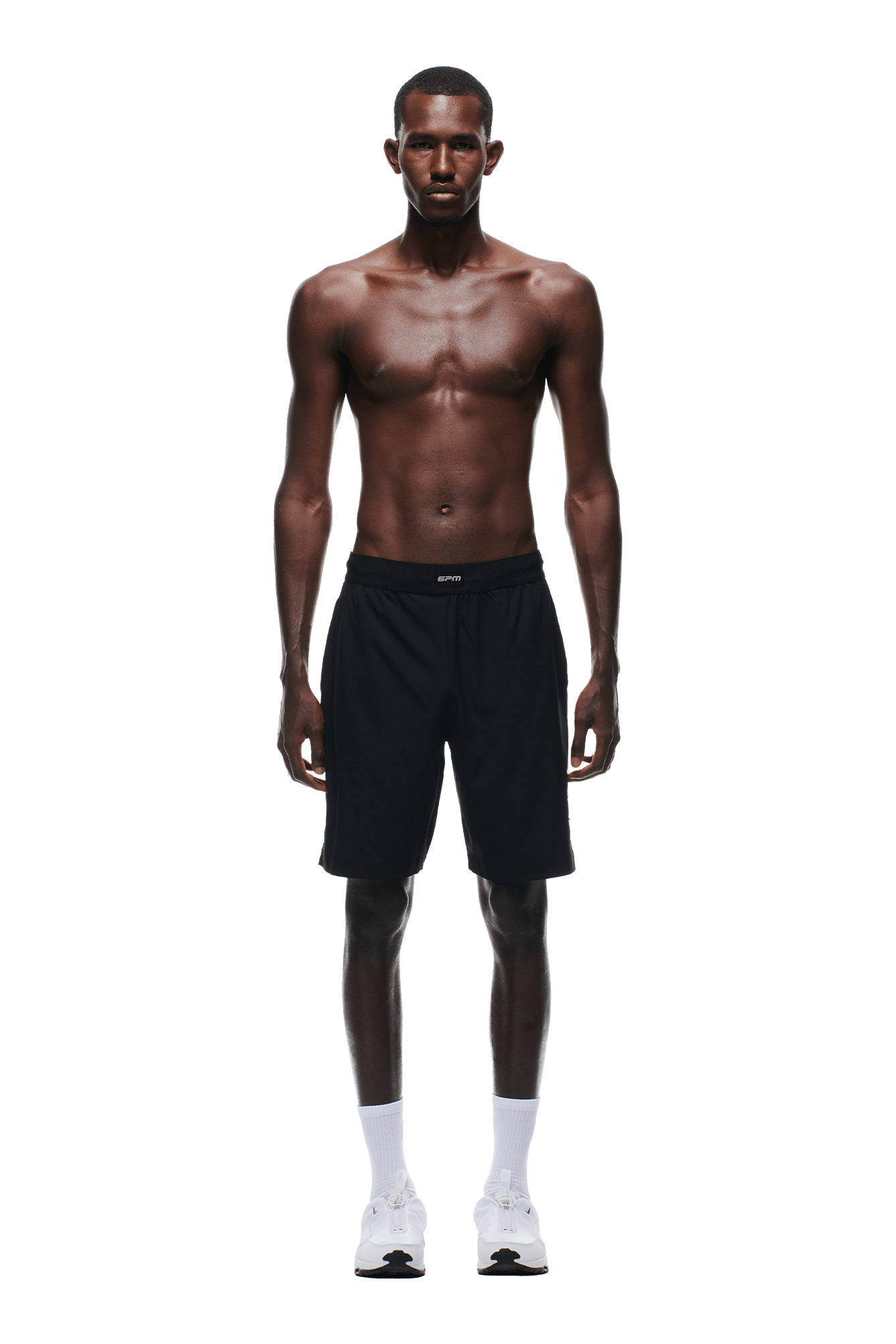 A shirtless man faces forward, wearing 6PM SPORTS SHORTS BLACK by 6PM, along with white socks and white sneakers, against a plain green background.