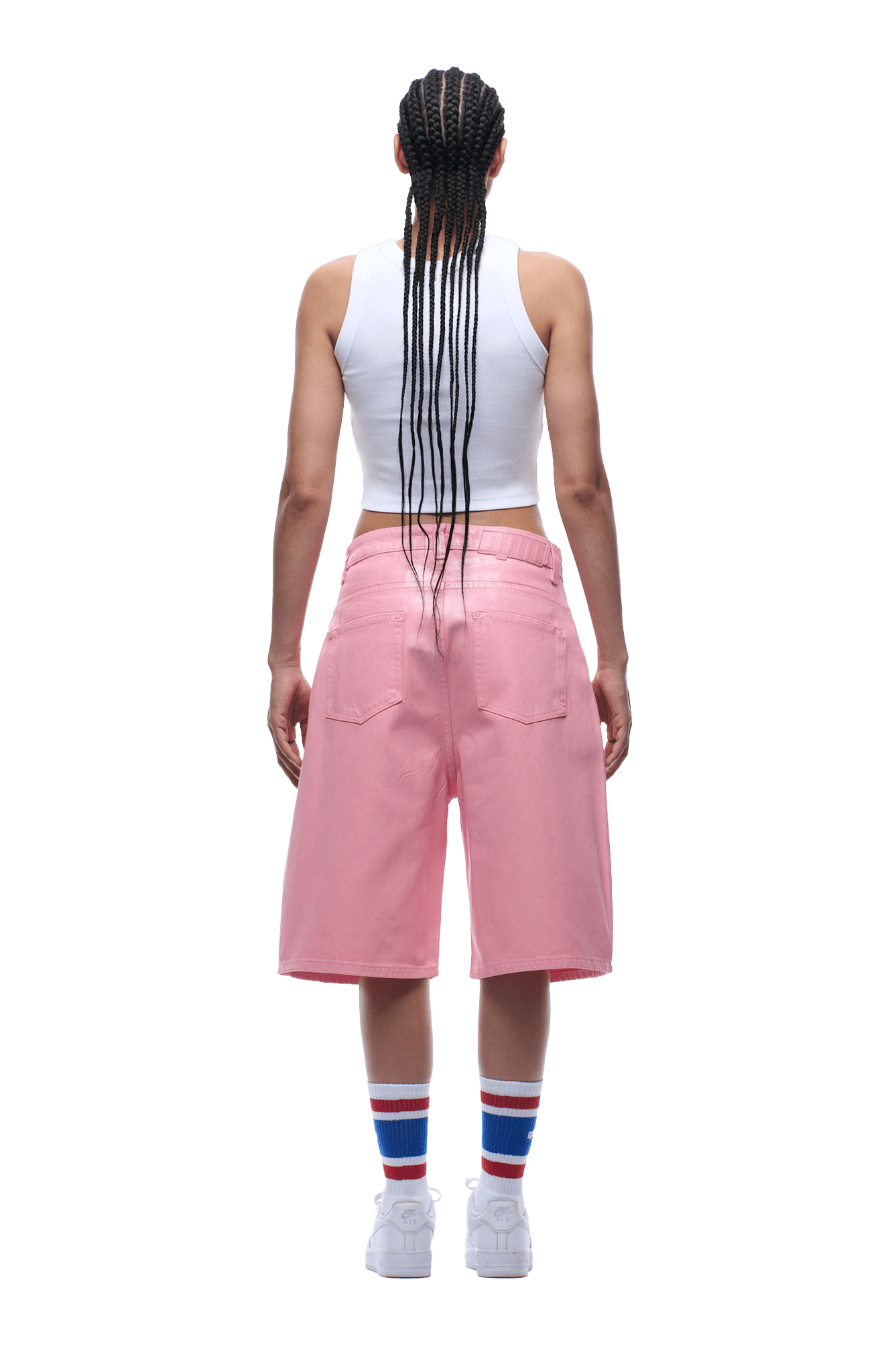 A person with long braids, wearing a white sleeveless top, 6PM COATED DENIM SHORTS PINK, white sneakers, and white socks with blue and red stripes stands facing away from the camera on a green background.