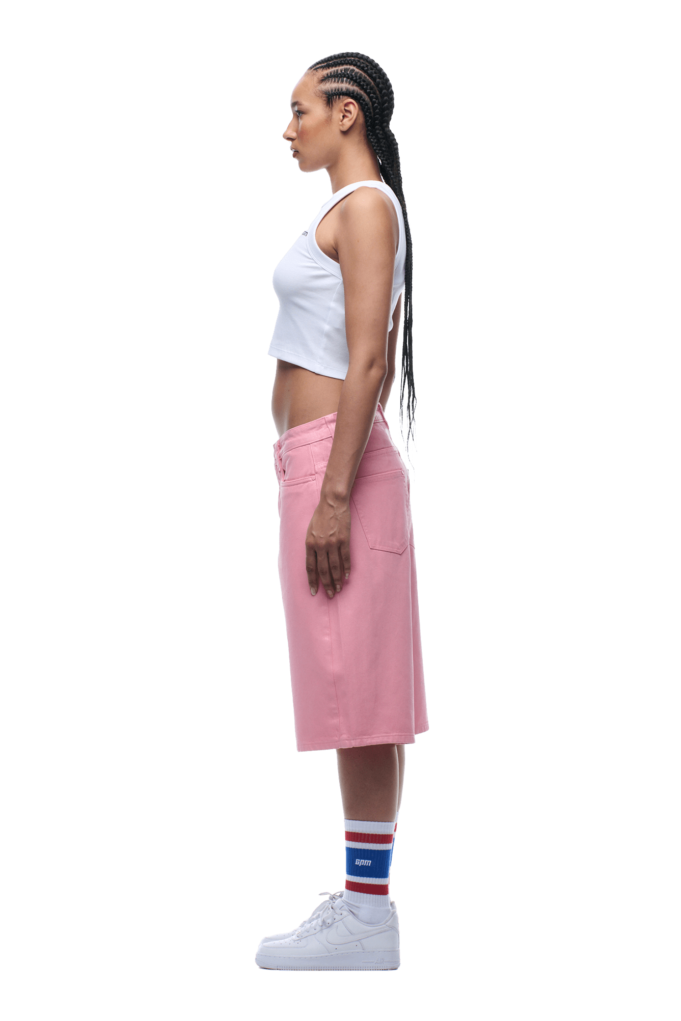 A woman stands in profile with long braided hair, wearing 6PM’s COATED DENIM SHORTS PINK, a white crop top, striped socks, and white sneakers on a transparent background.