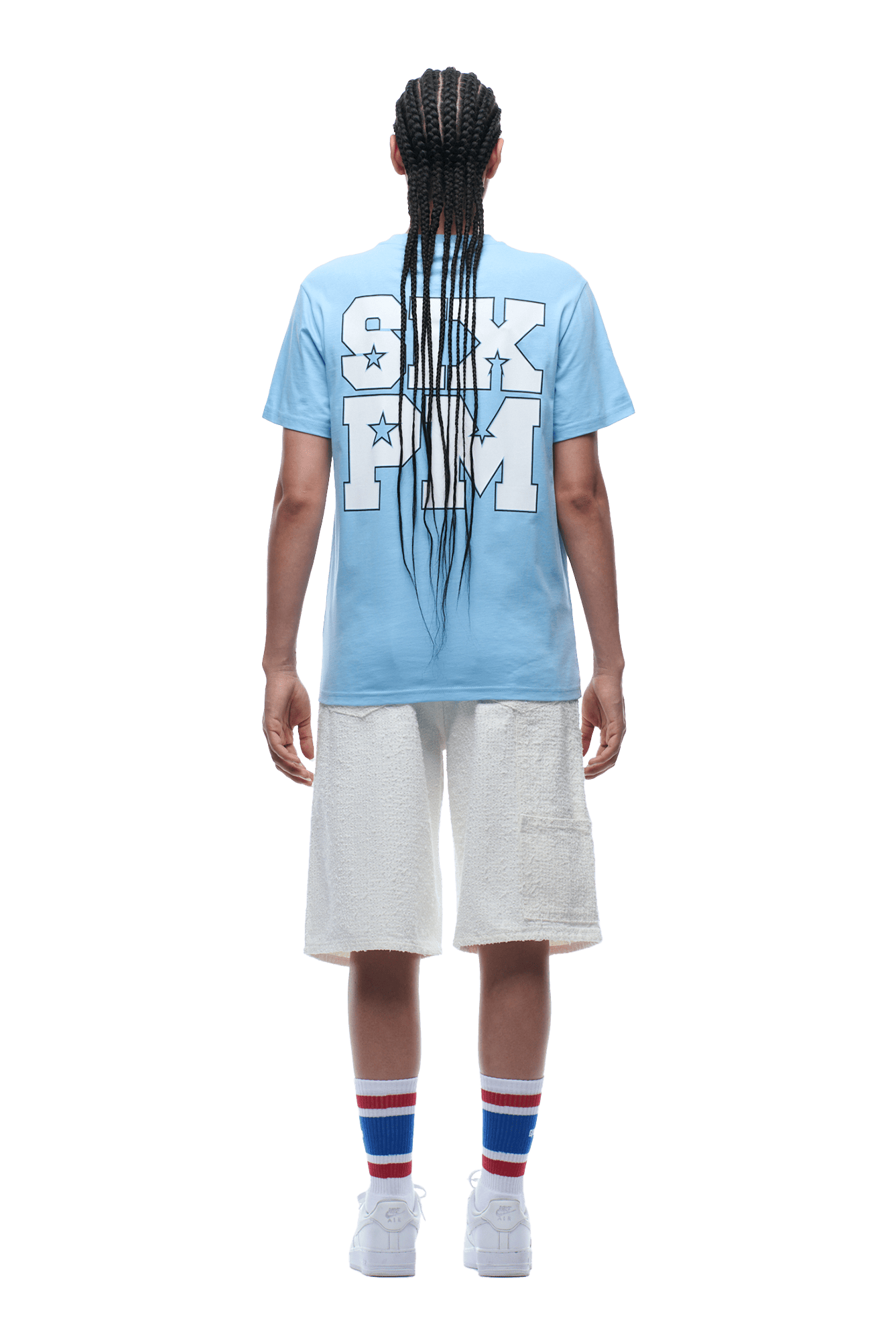 A person with long braids faces away, wearing the 6PM COLLEGE STAR TEE BLUE featuring bold back text, white shorts, striped socks, and white sneakers. The background is transparent.