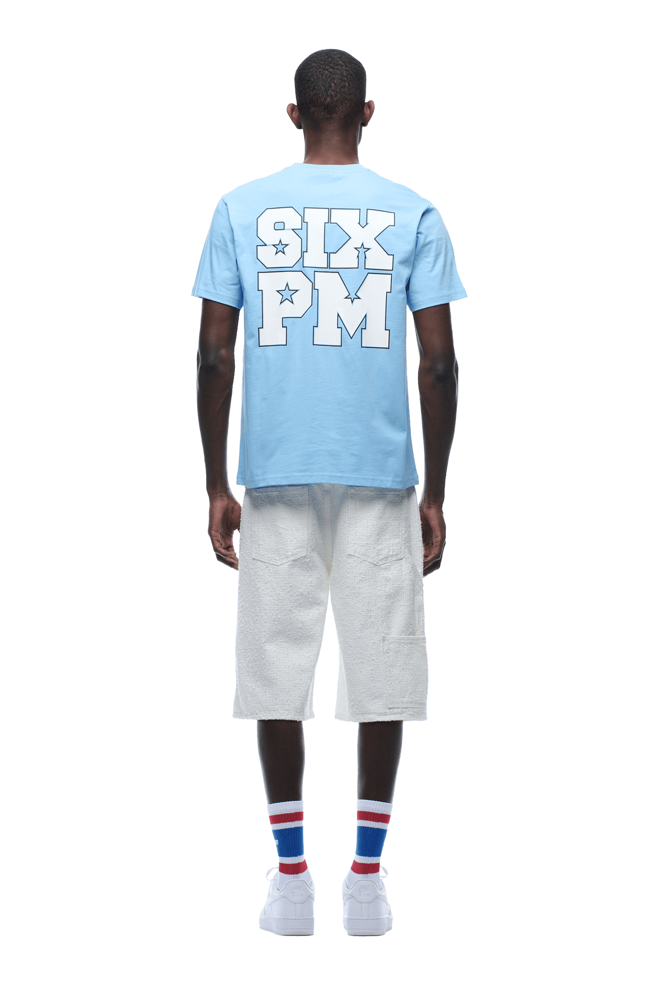 A person faces away, wearing the 6PM COLLEGE STAR TEE BLUE with bold white SIX PM lettering on the back, paired with white textured knee-length shorts, striped socks, and white sneakers. Background is transparent.