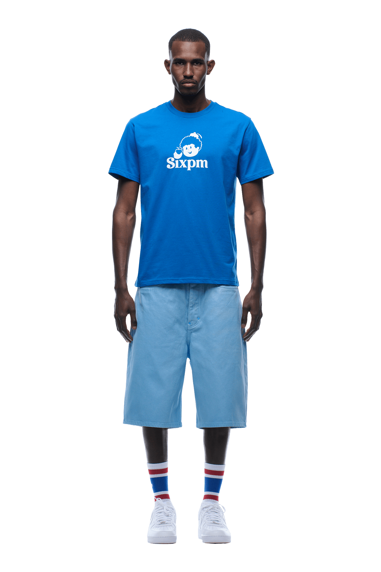 A man faces forward, wearing a blue APPLE BOY TEE BLUE by 6PM, light blue knee-length shorts, striped socks, and white sneakers on a transparent background.