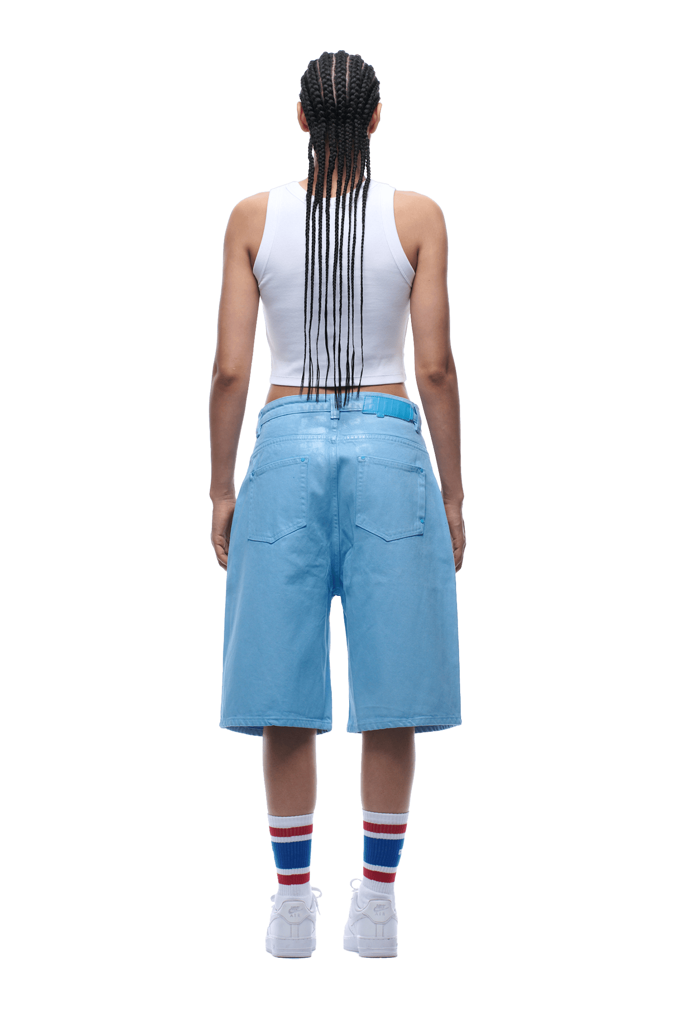 A person with long braids, in a white tank top, COATED DENIM SHORTS BABY BLUE, white socks with red and blue stripes, and white sneakers stands facing away from the camera on a transparent background.