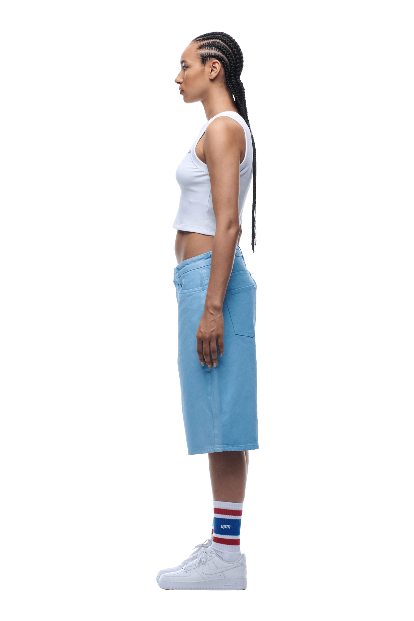 A woman with braided hair stands in profile against a plain background wearing COATED DENIM SHORTS BABY BLUE, a white crop top, white sneakers, and striped socks.