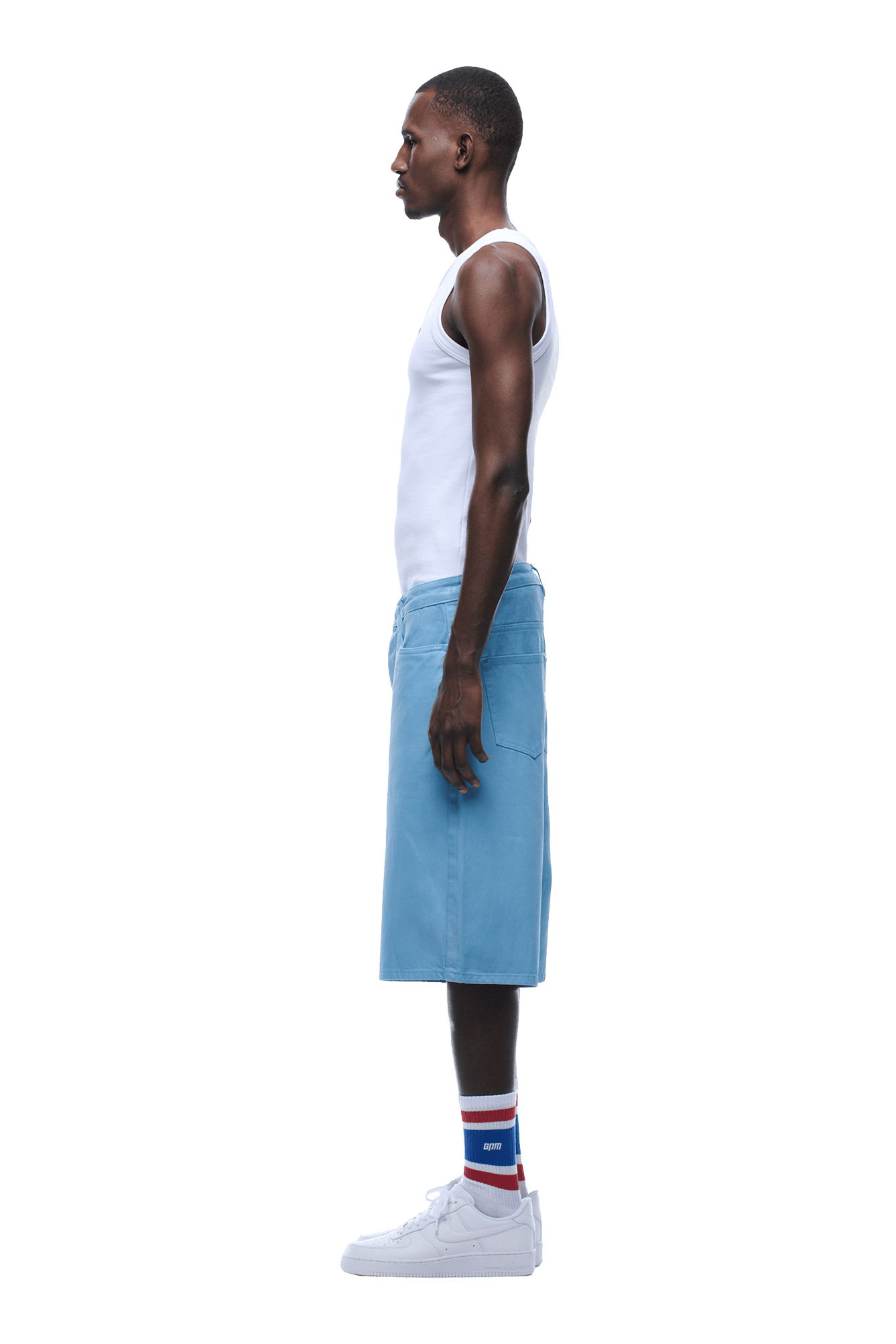 A man stands in profile wearing a white tank top, COATED DENIM SHORTS BABY BLUE, striped socks, and white sneakers against a transparent background.