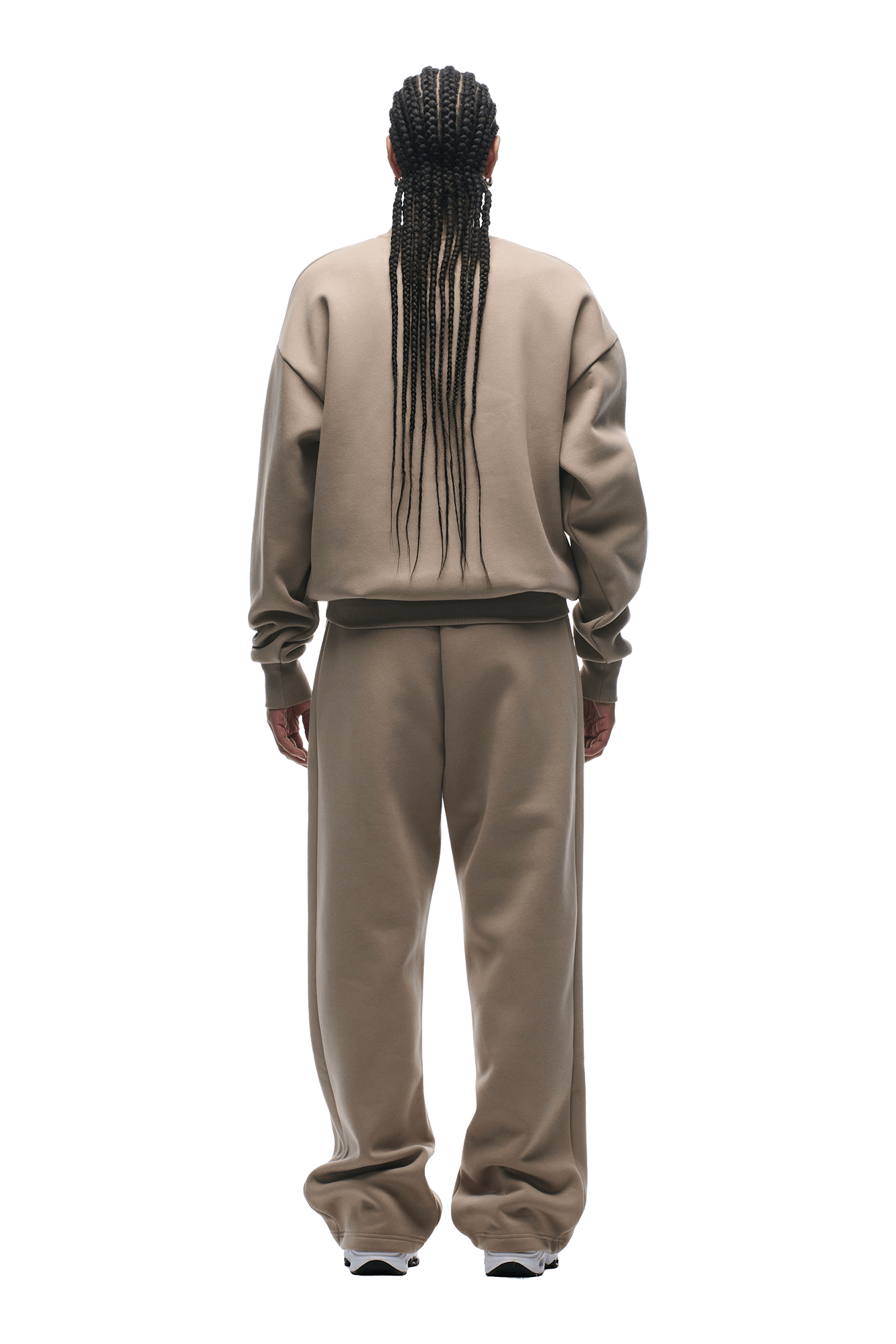 A person with long braids, wearing 6PMs CREWNECK MOCCA CREAM— a boxy-fit taupe sweatshirt with ribbed trims and matching pants— stands facing away on a transparent background.