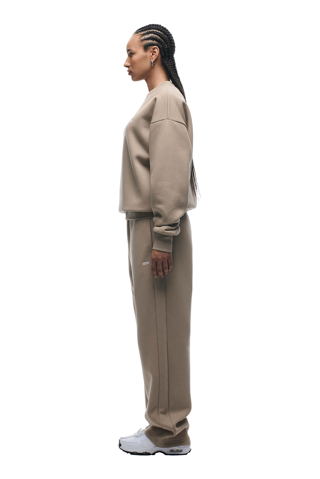 A woman with braided hair stands in profile, wearing the 6PM CREWNECK MOCCA CREAM sweatshirt and matching sweatpants with white sneakers, set against a plain green background.