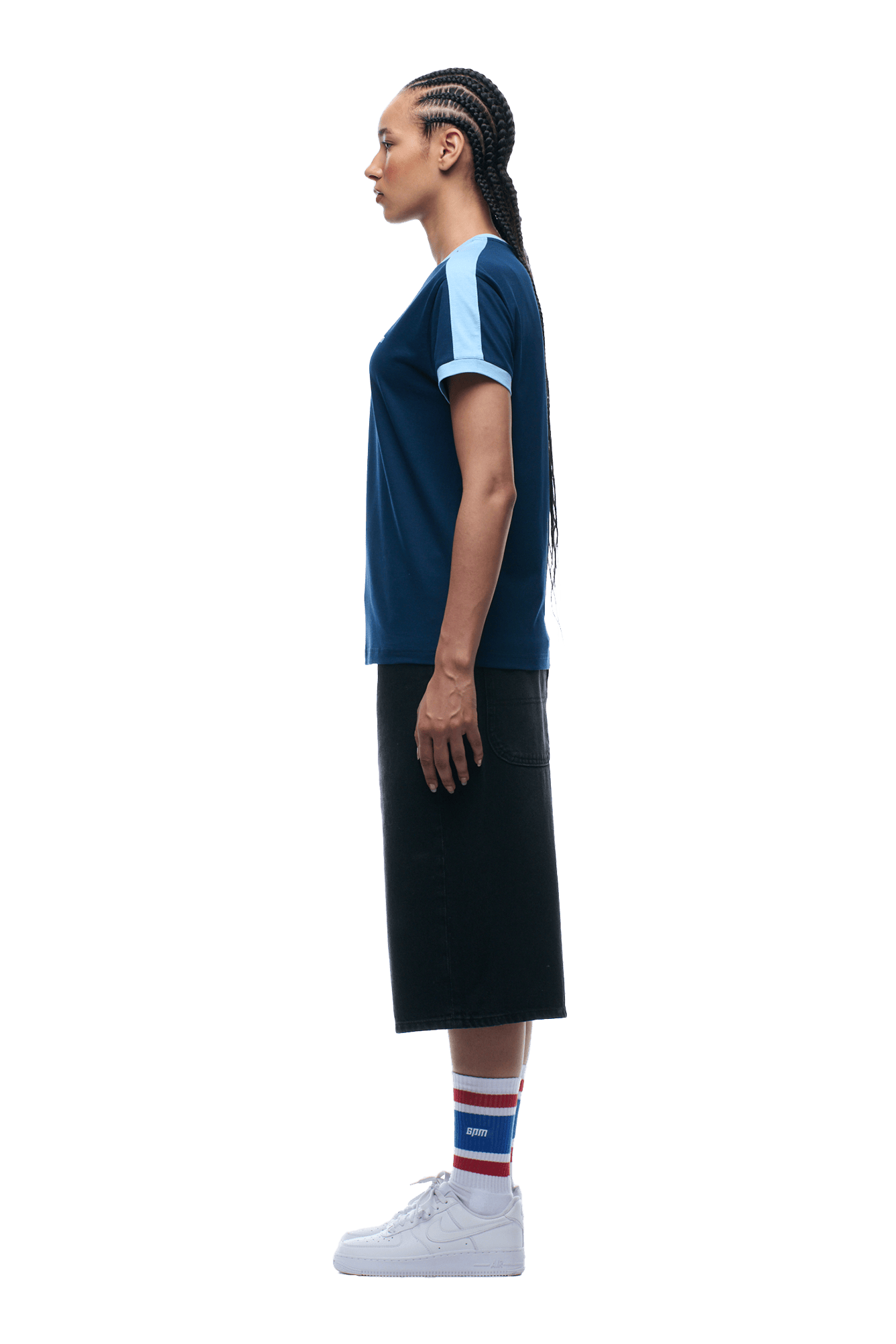 A woman with braided hair stands in profile on a transparent background, wearing the 6PM SLIM FIT RINGER TEE NAVY, a black midi skirt, white sneakers, and colorful striped socks.