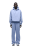 A person wears the HOODIE SOFT VIOLET, an oversized light blue raglan-sleeved hoodie with a kangaroo pocket and matching wide-leg pants, paired with white athletic shoes. Suitable for everyday use. Transparent background.