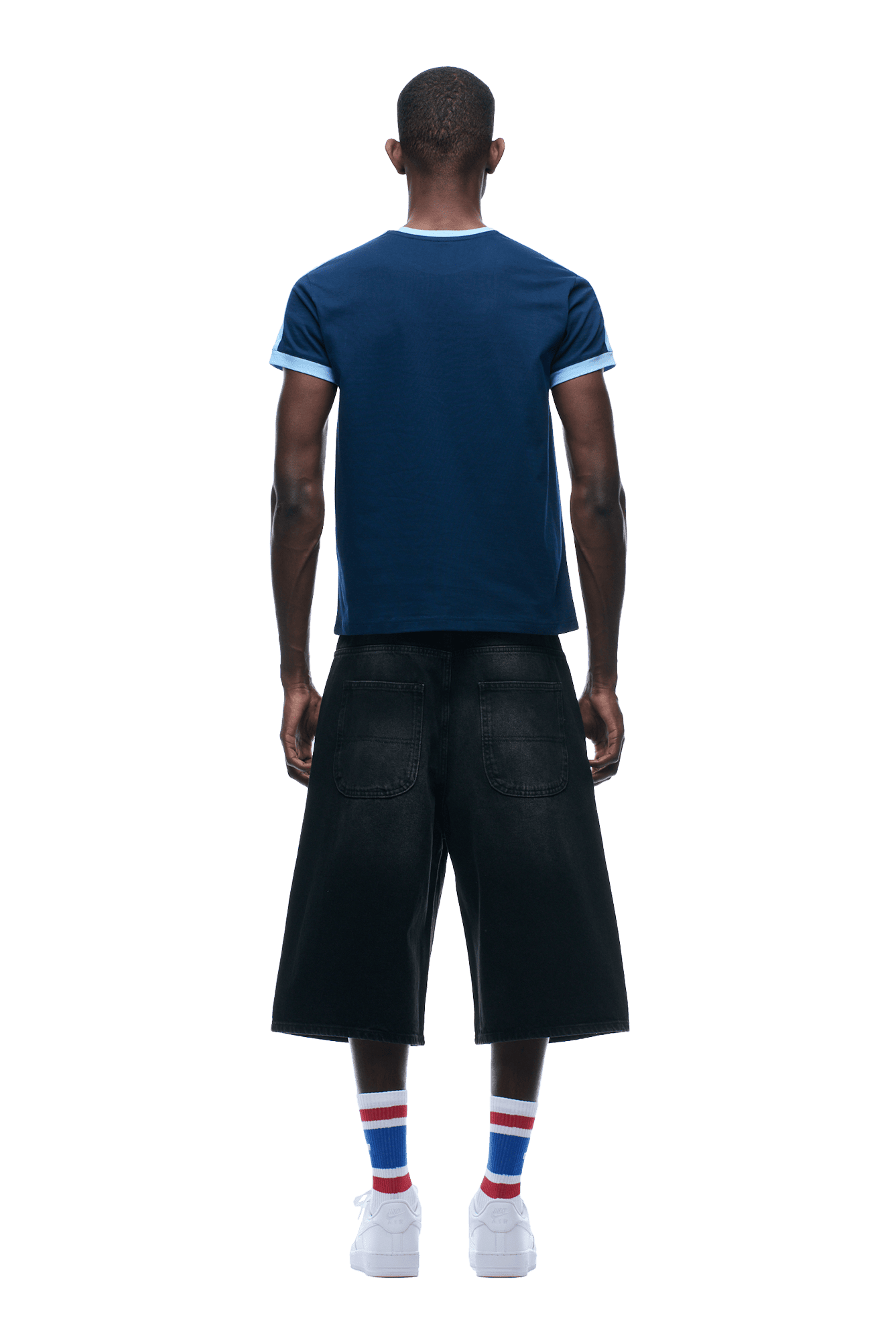 A person stands on a plain green background, facing away and wearing the 6PM SLIM FIT RINGER TEE NAVY, paired with black wide-leg denim shorts, white socks with blue and red stripes, and white sneakers.