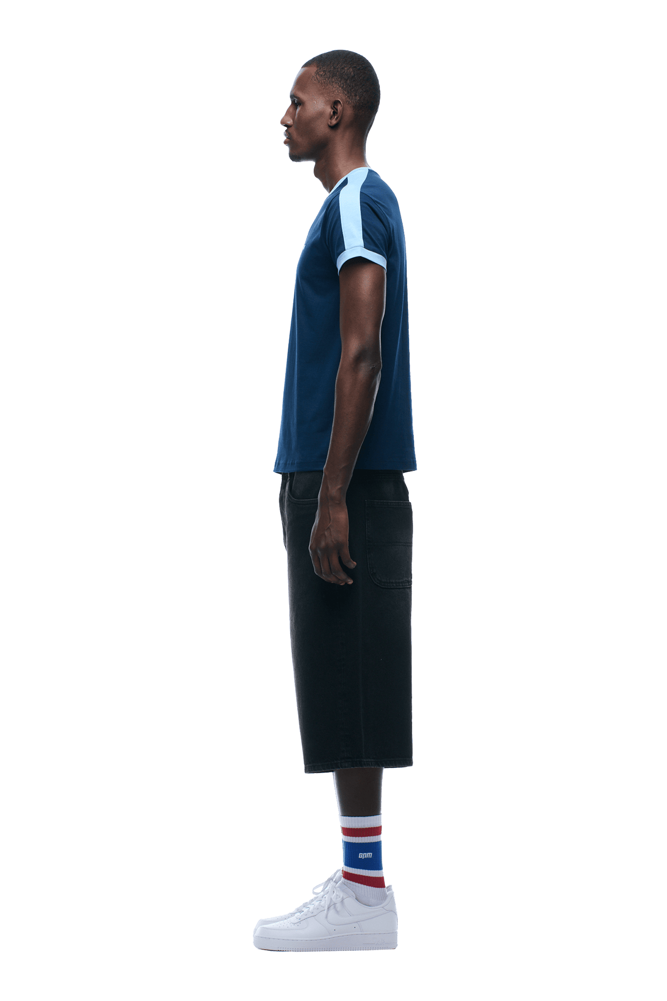A man in profile faces left, wearing the SLIM FIT RINGER TEE NAVY by 6PM with light blue sleeves, black knee-length shorts, white sneakers, and striped socks. The background is transparent.