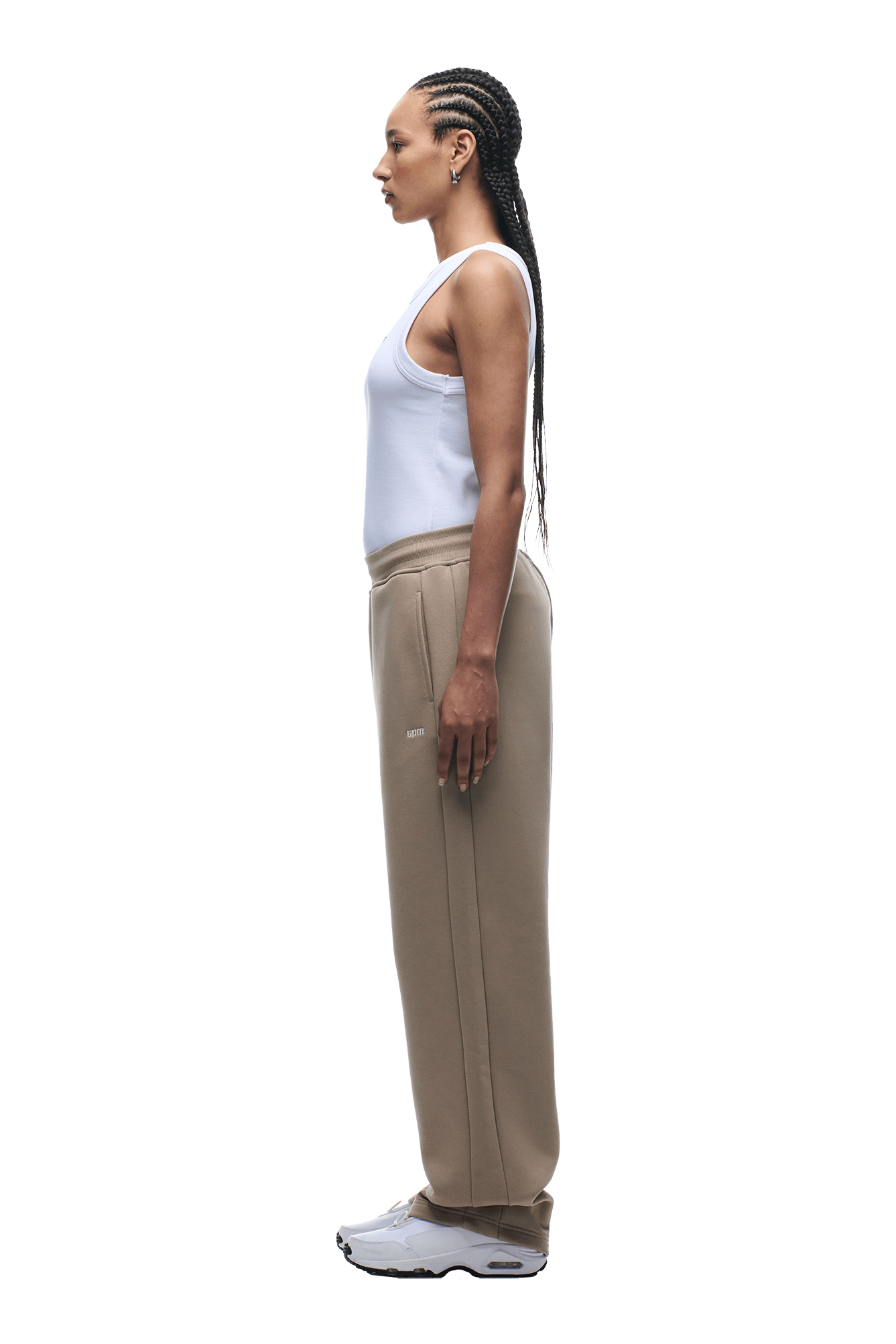 A person with braided hair stands in profile, facing left against a plain background, wearing a white sleeveless top, 6PMs OPEN LEG JOGGER MOCCA CREAM sweatpants, and white sneakers—a modern classic look.