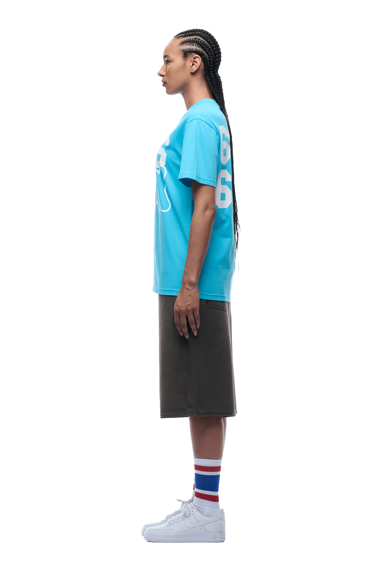 A woman in profile with braided hair wears the 6PM 660660 TEE BLUE, a knee-length dark skirt, white athletic shoes, and white socks with red and blue stripes, posed against a plain background.
