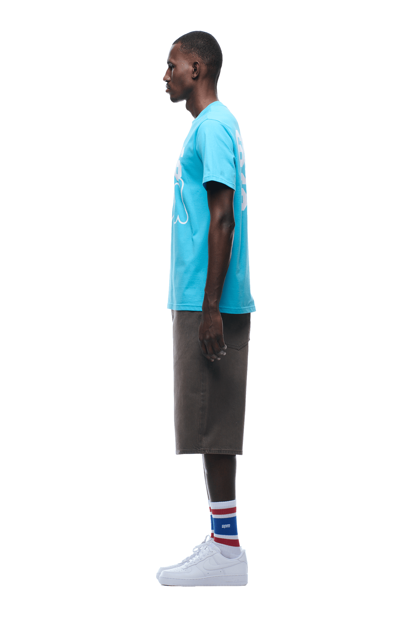 A man stands in profile facing left, wearing a 6PM 660660 TEE BLUE, gray knee-length shorts, blue-red-white striped socks, and white sneakers on a transparent background.