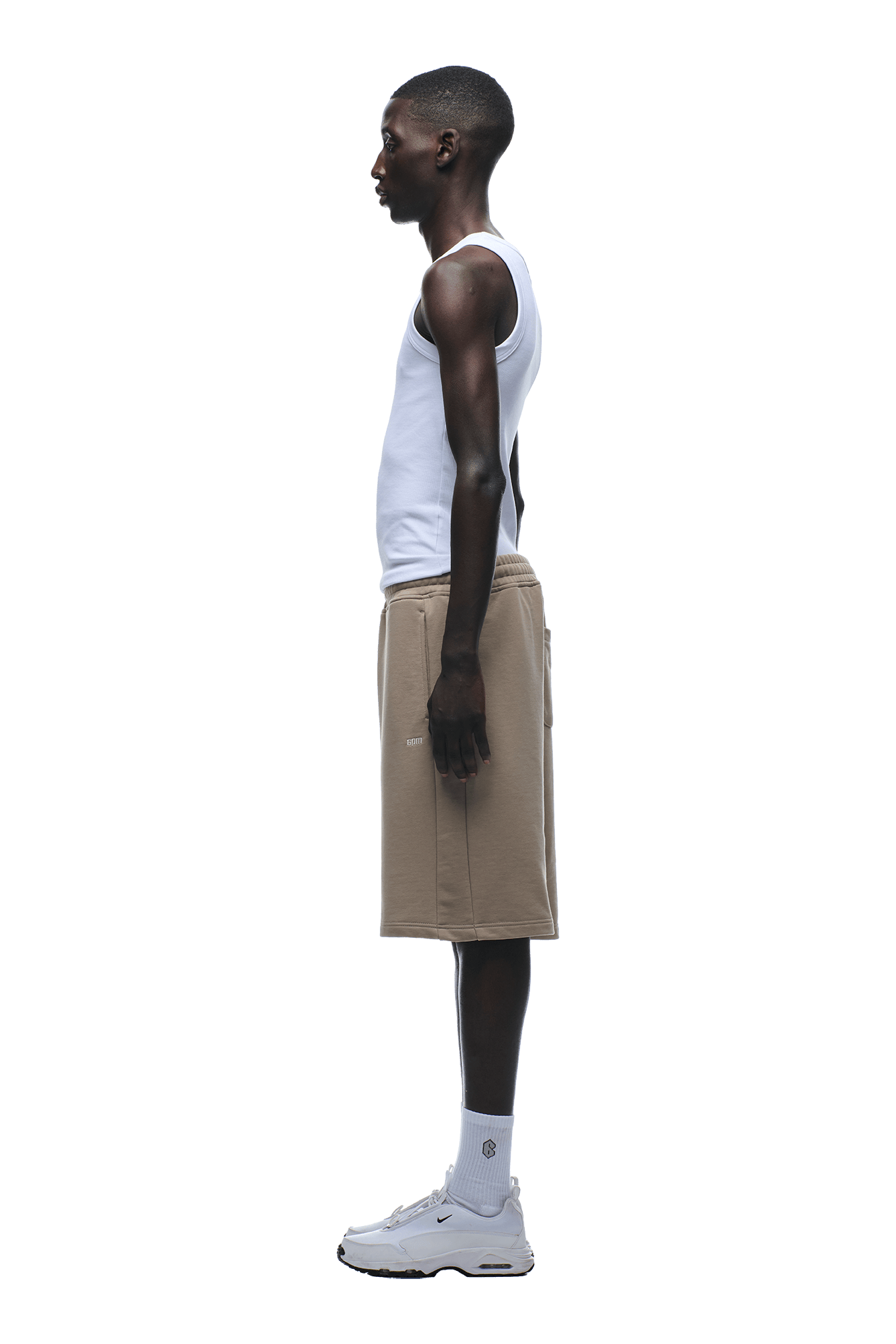 A man in profile faces left, wearing a white tank top, 6PM+ SHORTS MOCCA CREAM by 6PM—relaxed fit beige shorts with a drawstring waist—paired with white socks and sneakers against a plain background.