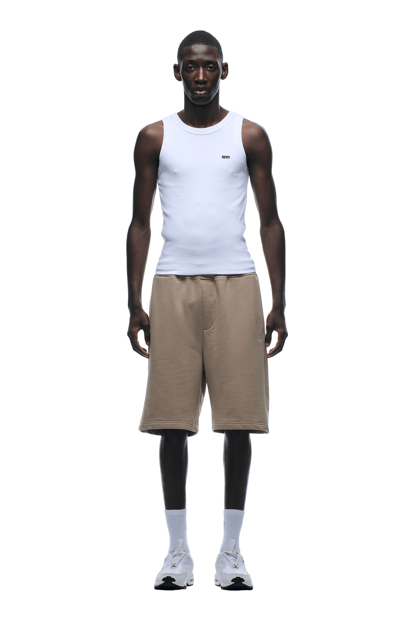 A man stands facing forward against a plain green background, wearing 6PM 6PM+ SHORTS MOCCA CREAM paired with a white sleeveless tank top, white crew socks, and sneakers—perfect for lounging.