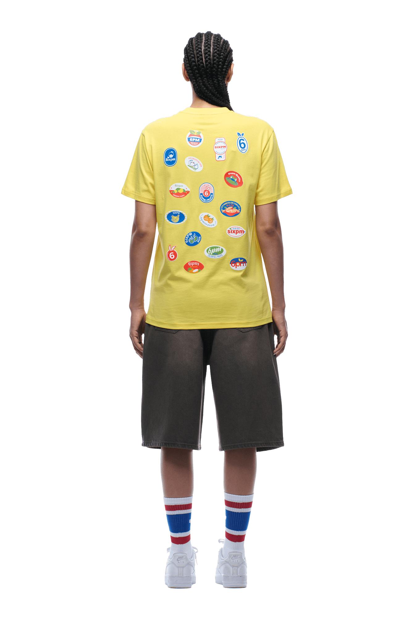 Person facing away, wearing the 6PM BANANA STICKER TEE YELLOW with colorful logos on the back, dark knee-length shorts, white sneakers, and tall white socks with blue and red stripes. Transparent background.
