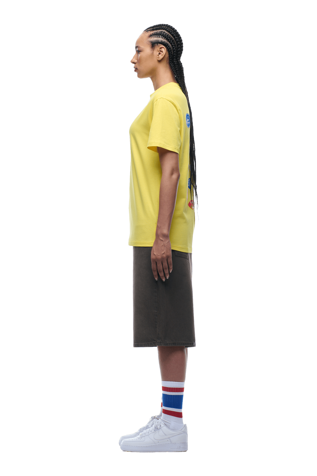 A person with long braids stands in profile, wearing the 6PM BANANA STICKER TEE YELLOW, a dark knee-length skirt, striped socks, and white sneakers against a plain background.