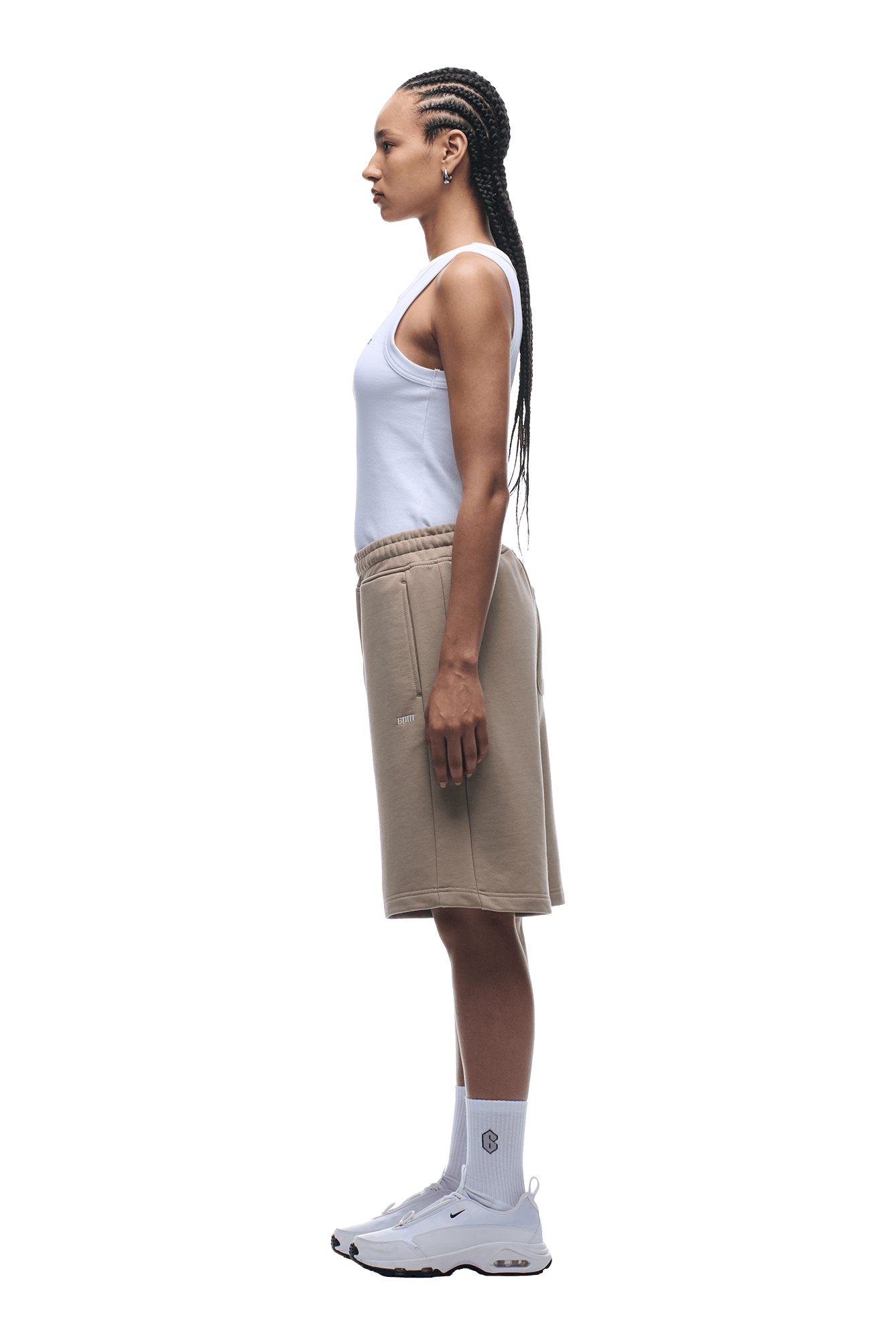 A woman with long braided hair stands in profile, wearing 6PMs 6PM+ SHORTS MOCCA CREAM—beige, knee-length shorts with a drawstring waist—paired with a white tank top, crew socks, and sneakers. The background is transparent.