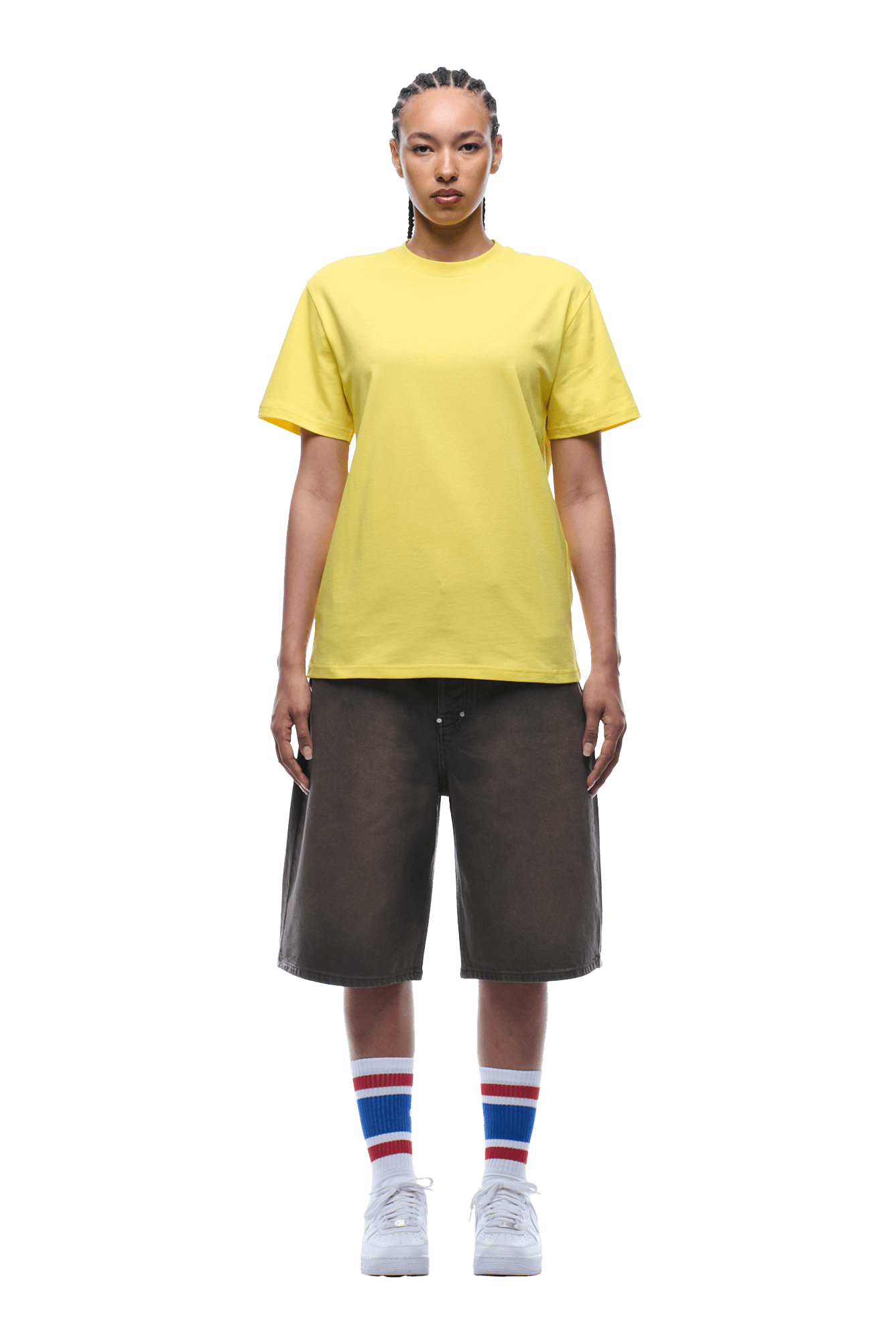A person with braided hair faces forward, wearing the 6PM BANANA STICKER TEE YELLOW, loose dark shorts, white sneakers, and blue-red-white striped crew socks, set against a transparent background.