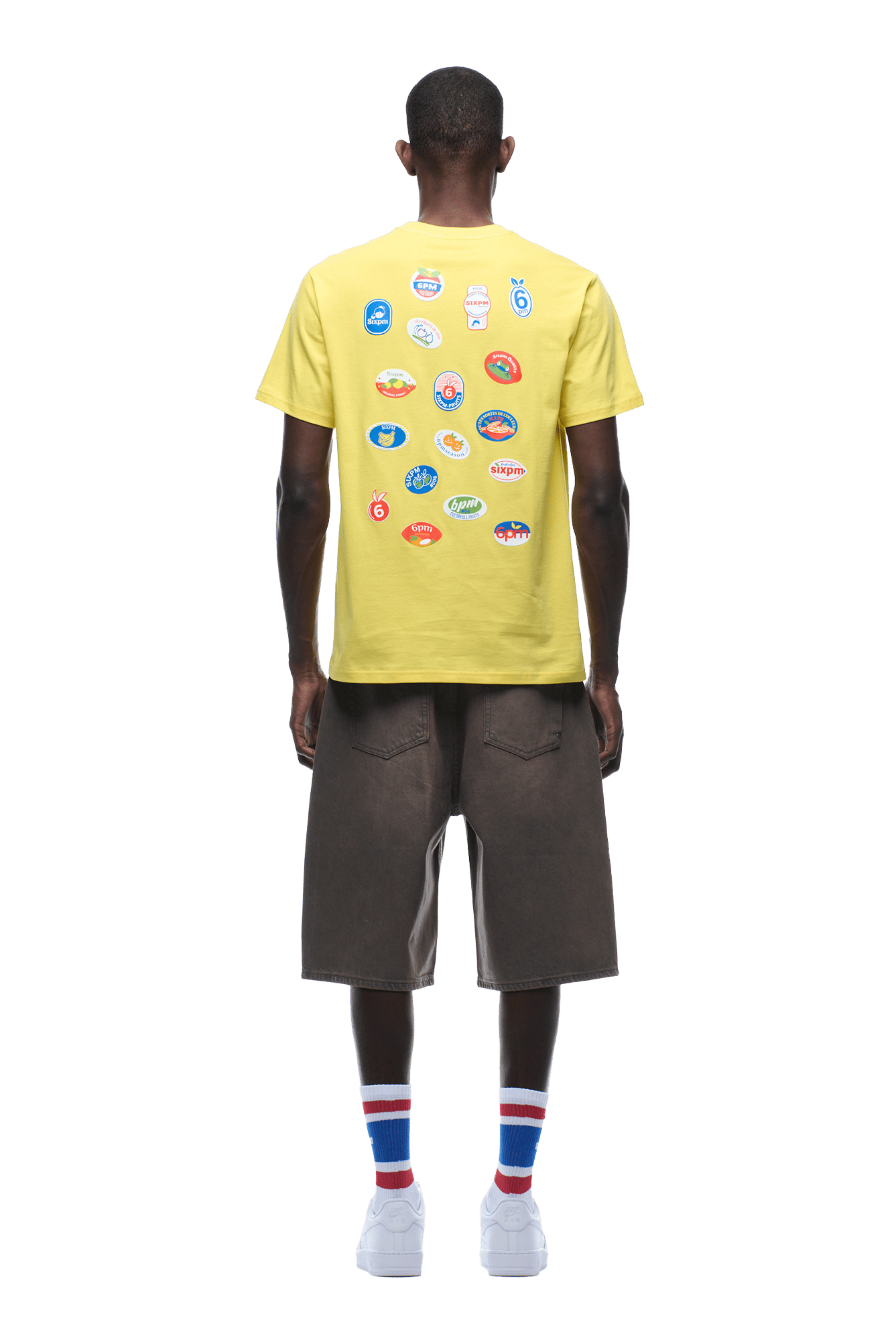 A man, facing away on a transparent background, wears the 6PM BANANA STICKER TEE YELLOW featuring colorful back graphic patches, brown knee-length shorts, white sneakers, and tall white socks with red and blue stripes.
