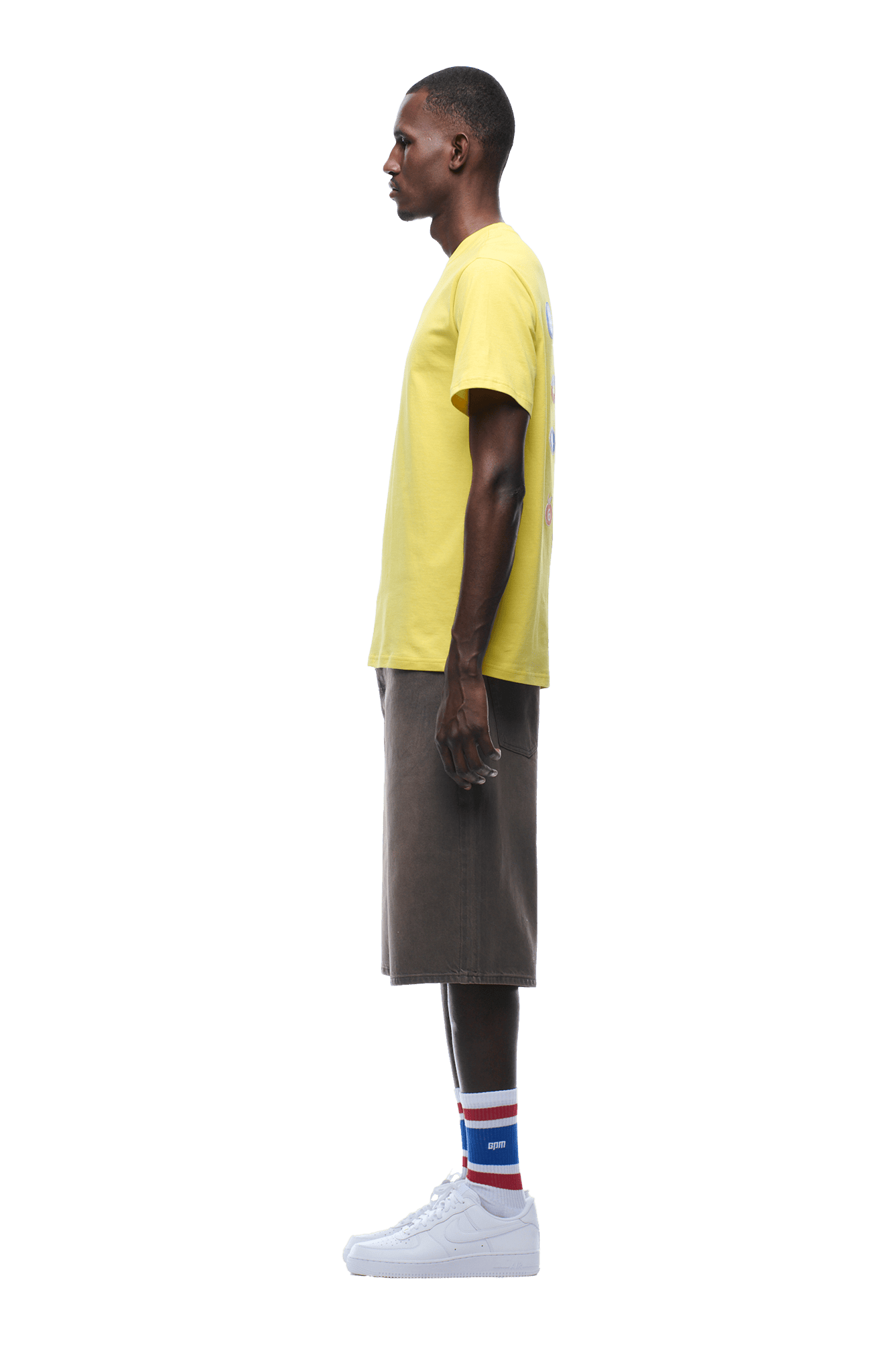 A man in profile facing left wears the 6PM BANANA STICKER TEE YELLOW, paired with gray knee-length shorts, white sneakers, and white socks with red and blue stripes against a plain background.