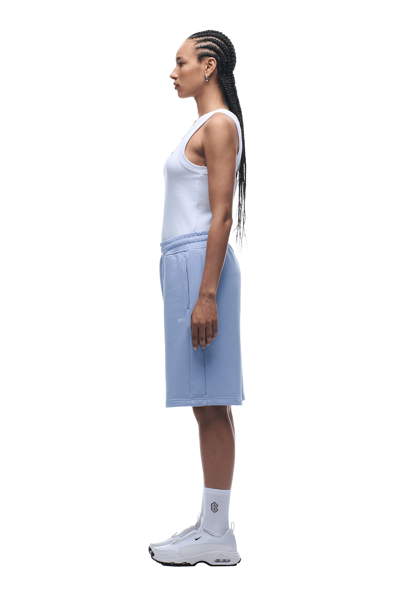 A woman with long braided hair stands in profile, lounging in a white tank top and 6PM+ SHORTS SOFT VIOLET with a drawstring waist, paired with white socks and sneakers, against a plain background.