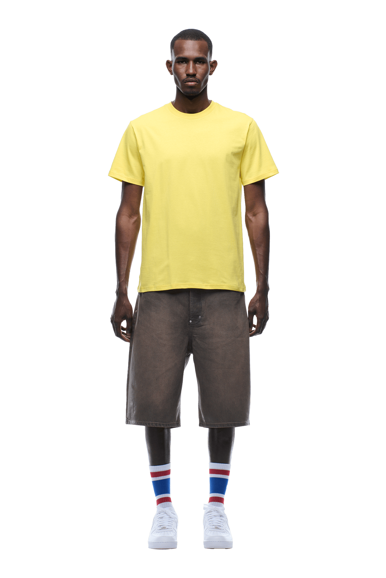 A man stands facing forward wearing the 6PM BANANA STICKER TEE YELLOW, loose gray shorts, white sneakers, and white socks with blue and red stripes. The background is transparent.