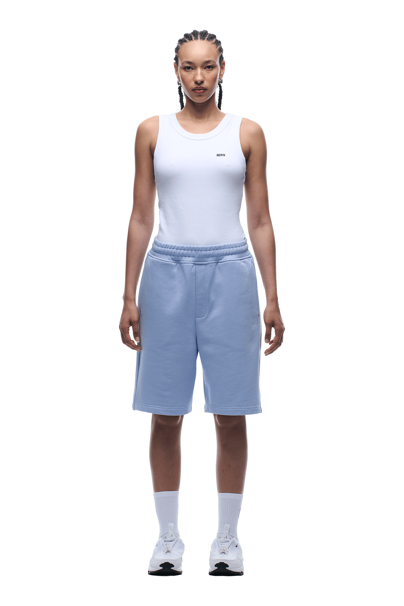 A person faces forward, wearing a white sleeveless top and 6PM+ SHORTS SOFT VIOLET—light blue knee-length shorts with a relaxed fit and drawstring waist—paired with white crew socks, white sneakers, and braided hair, against a plain background.