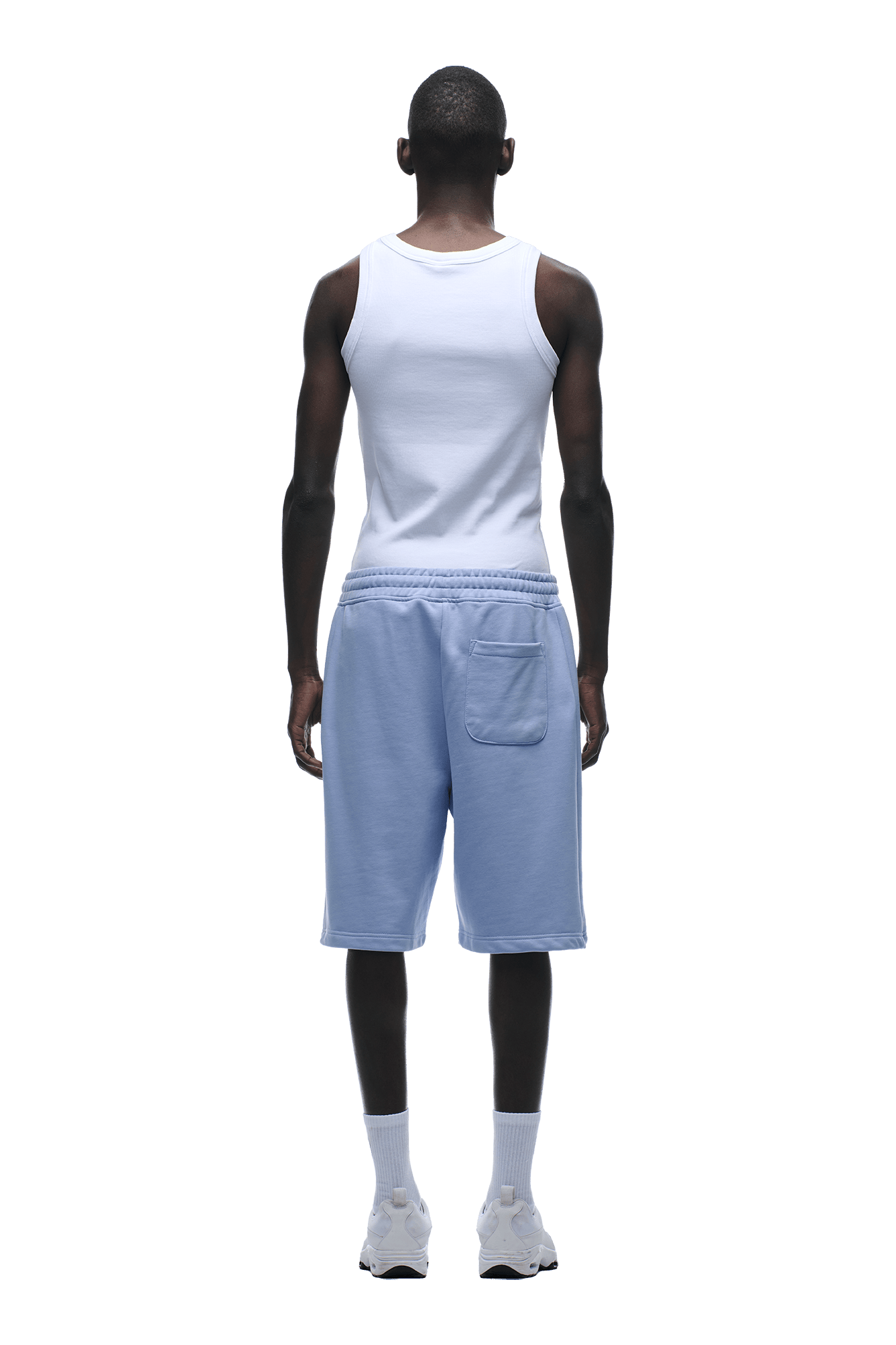 A person stands facing away, wearing a white tank top and 6PM+ SHORTS SOFT VIOLET—light blue, relaxed-fit, knee-length shorts with a drawstring waist and back pocket—plus white socks and sneakers, against a plain green background.