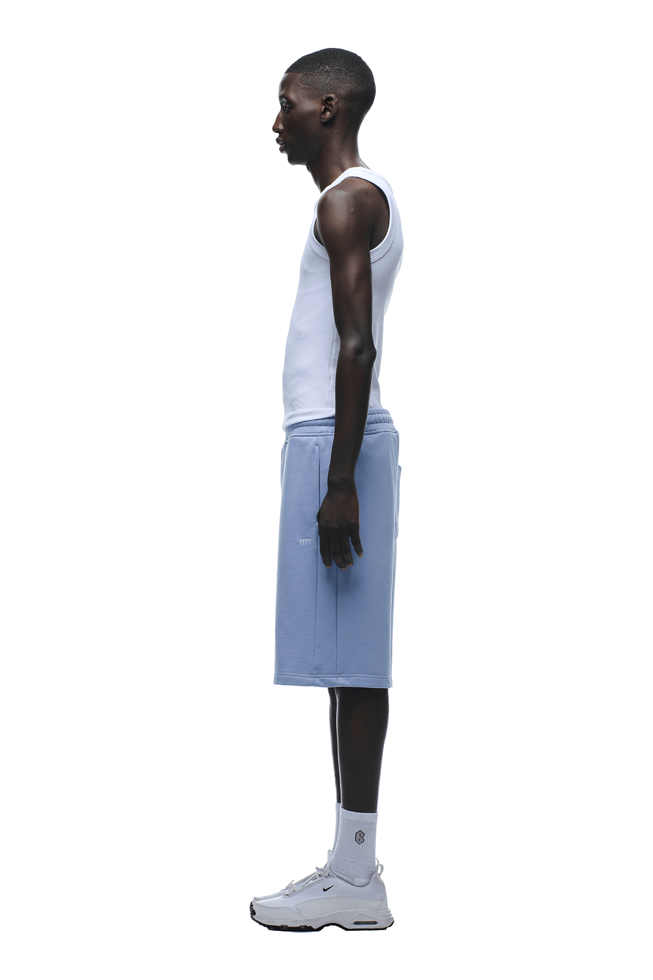 A man stands in profile view wearing a white tank top, 6PM+ SHORTS SOFT VIOLET with a drawstring waist, white crew socks, and white sneakers against a plain background.