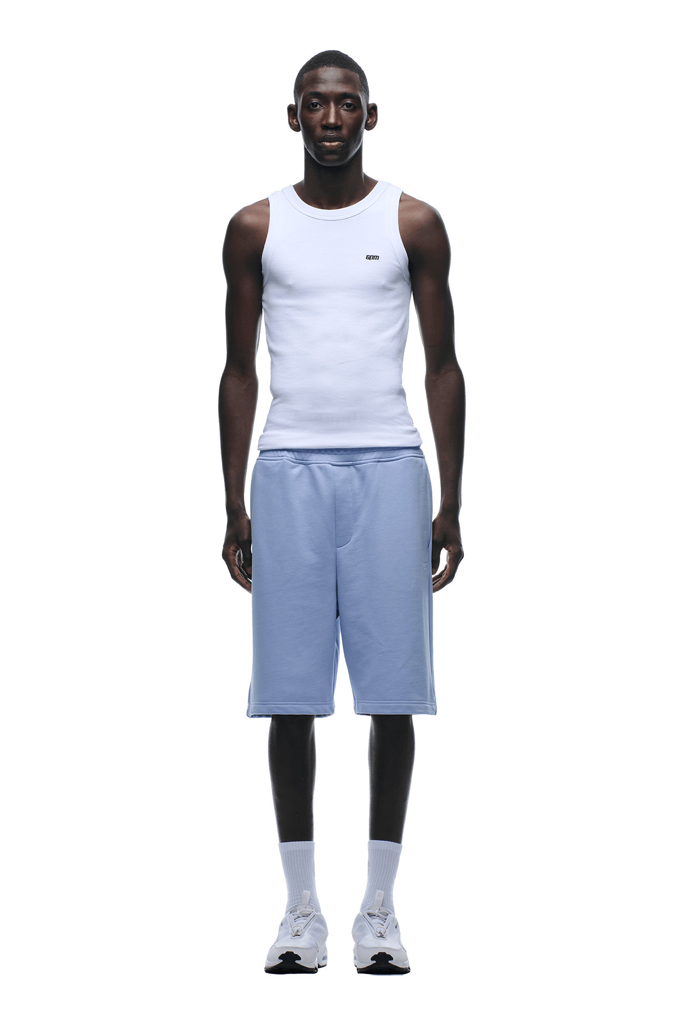 A man stands facing forward, wearing a fitted white tank top and 6PM+ SHORTS SOFT VIOLET, which are light blue, knee-length with a drawstring waist; he also wears white socks and sneakers. The background is transparent.