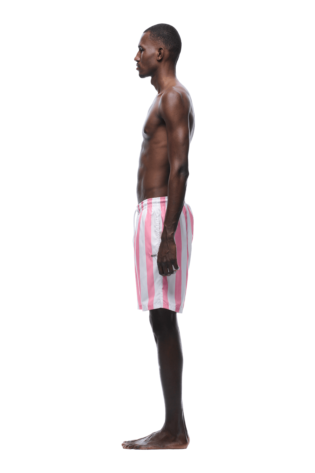 A young man stands in profile, facing left, barefoot and shirtless, wearing 6PM STRIPED SWIM SHORTS WHITE/PINK against a plain green background.