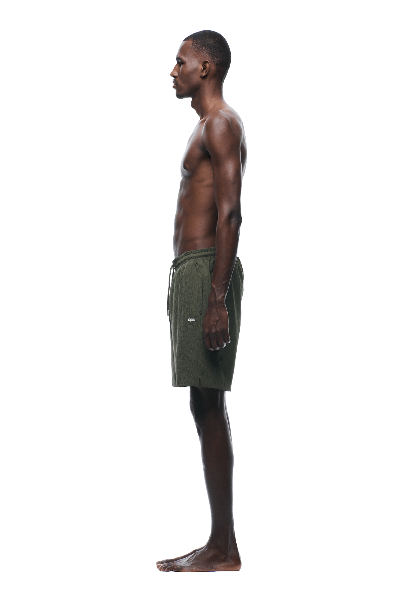 A man stands in profile facing left, barefoot, wearing 6PM SEERSUCKER SWIM SHORTS OLIVE against a plain green background.
