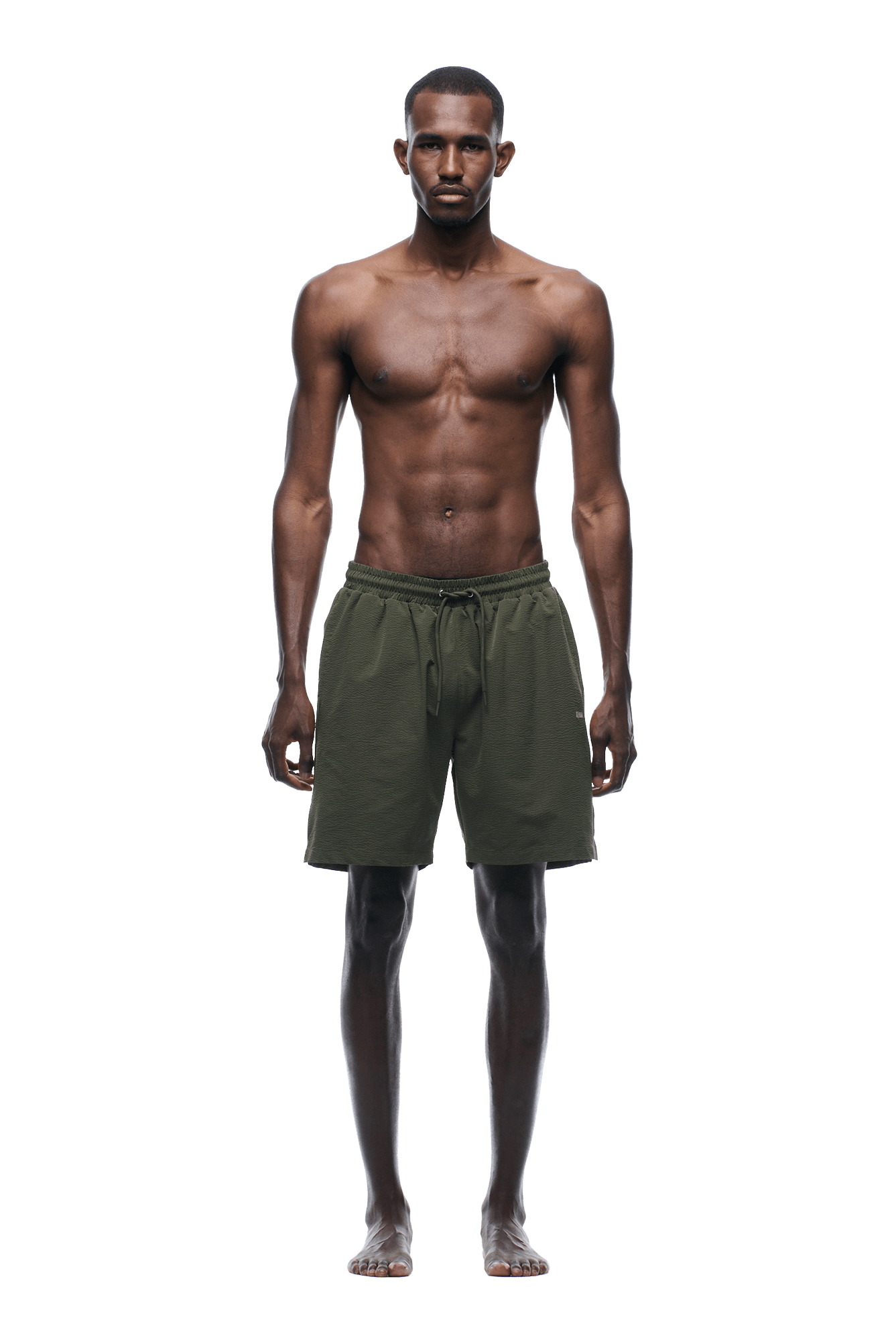 A man with a lean, muscular build stands barefoot facing forward, wearing 6PM SEERSUCKER SWIM SHORTS OLIVE. He is shirtless, arms relaxed at his sides, in front of a plain green background.