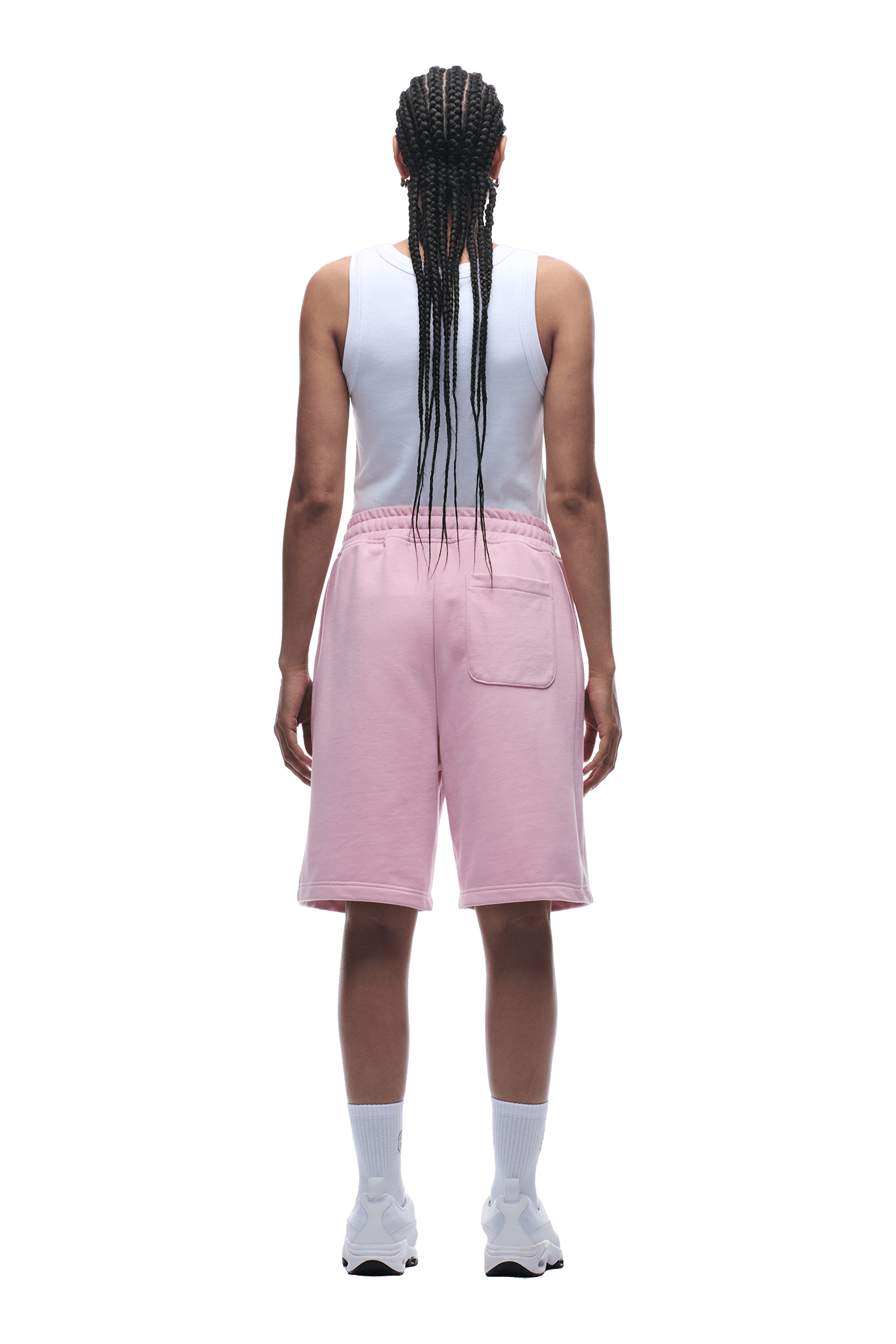 A person with long braids wears a white sleeveless top and 6PM 6PM+ SHORTS BABY PINK—relaxed fit, knee-length with a drawstring waist—plus white socks and sneakers, facing away from the camera on a plain background.