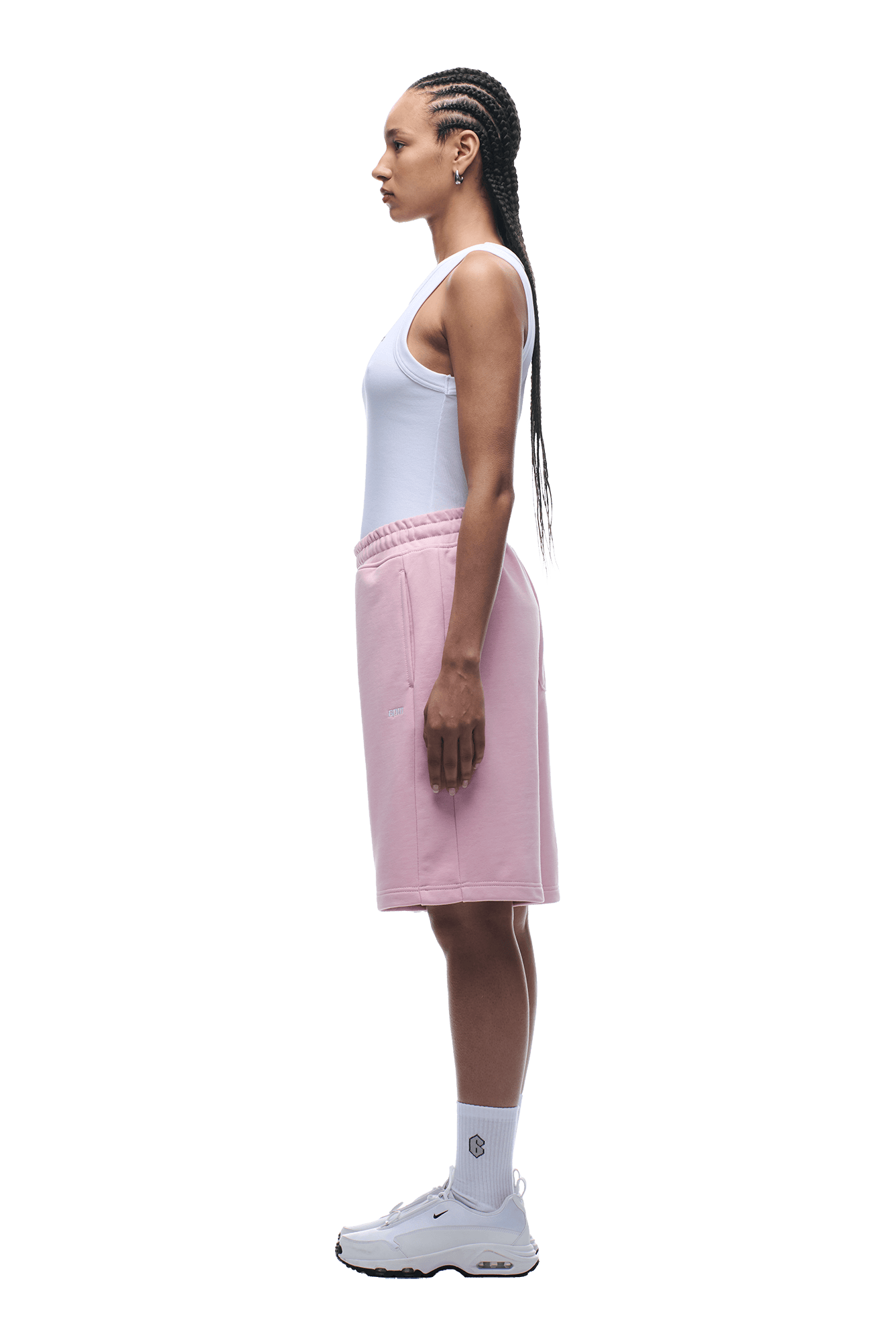 A woman in profile poses against a green backdrop, wearing 6PMs SHORTS BABY PINK with a drawstring waist, paired with a white tank top, white crew socks, and sneakers. Her braided hair drapes over her back for a relaxed look.