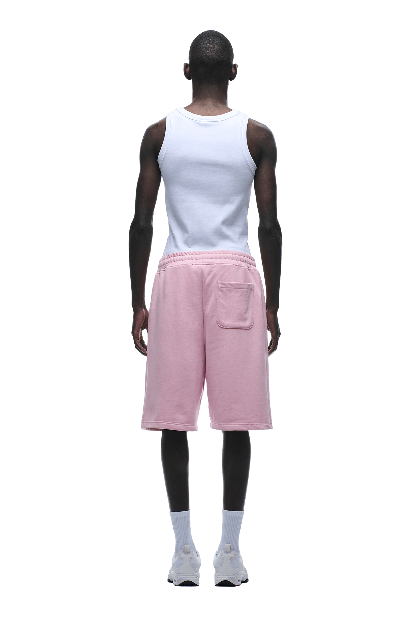 A person stands facing away, wearing a white tank top and 6PM’s relaxed fit 6PM+ SHORTS BABY PINK with a back pocket, white socks, and white sneakers, against a plain green background.
