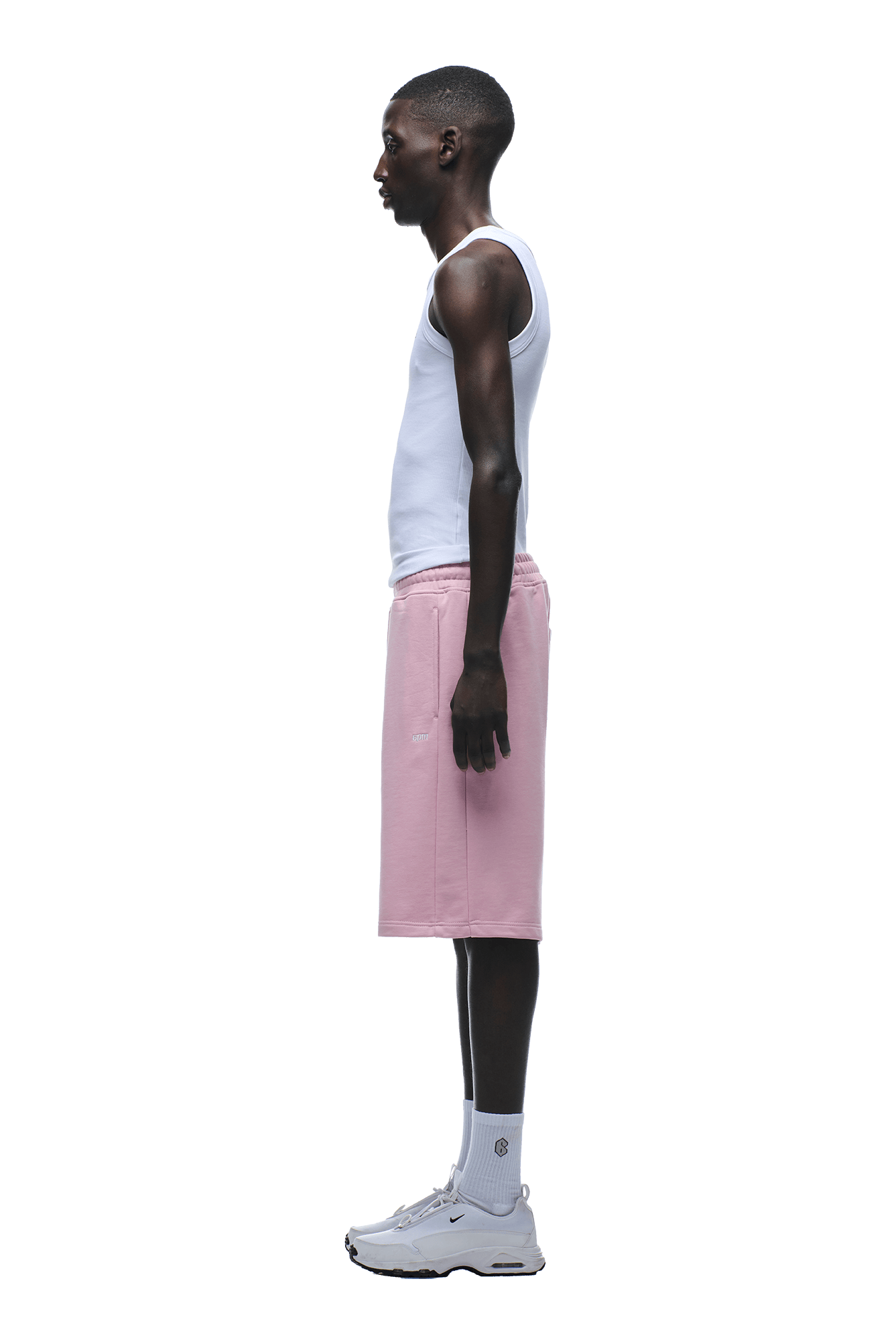 A man in profile wears the 6PM+ SHORTS BABY PINK by 6PM—relaxed fit, drawstring waist—styled with a white tank top, white socks, and sneakers against a plain background.