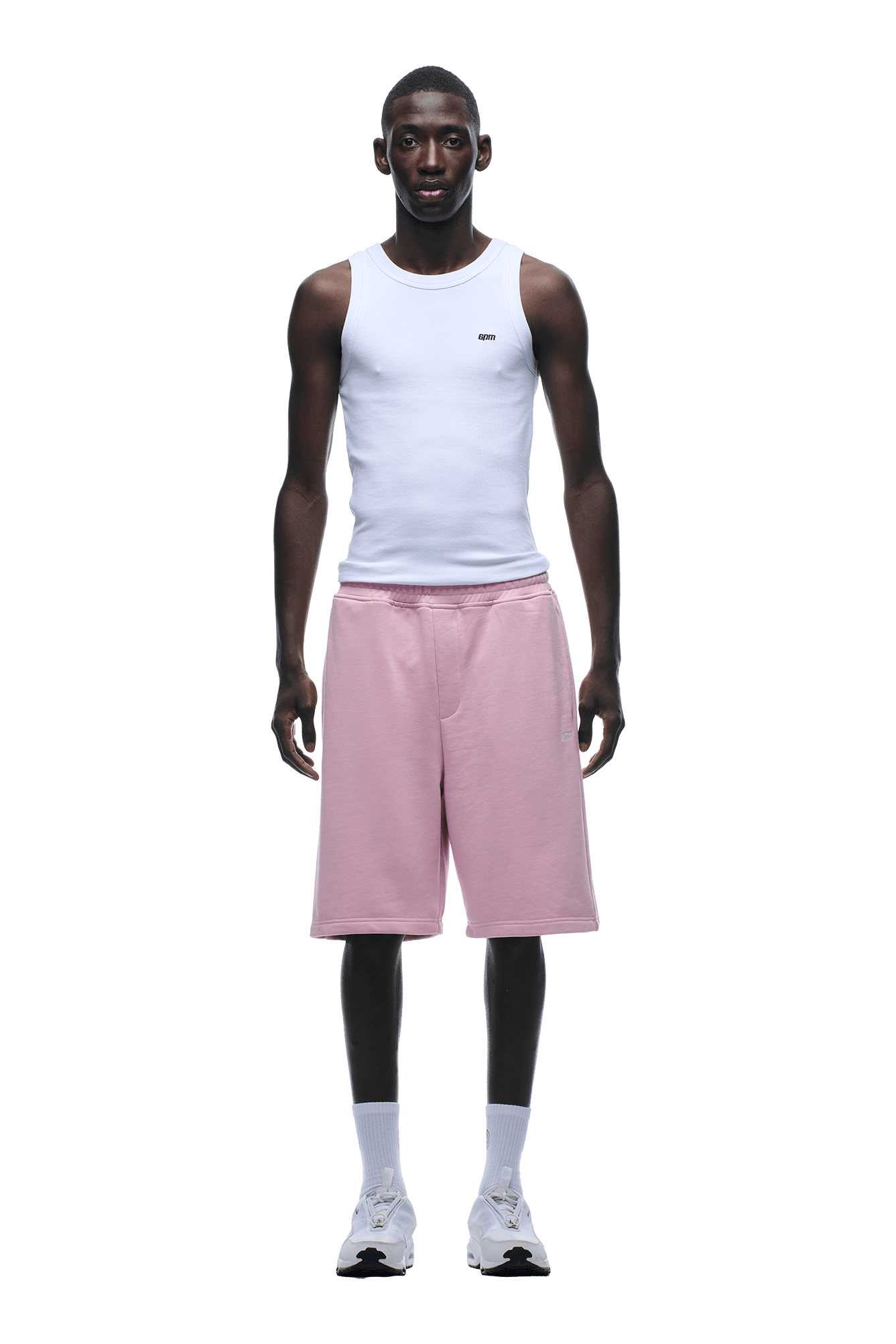 A man faces forward, wearing a white tank top and 6PM 6PM+ SHORTS BABY PINK—light pink, knee-length shorts with a relaxed fit and drawstring waist—paired with white crew socks and sneakers, against a plain background.