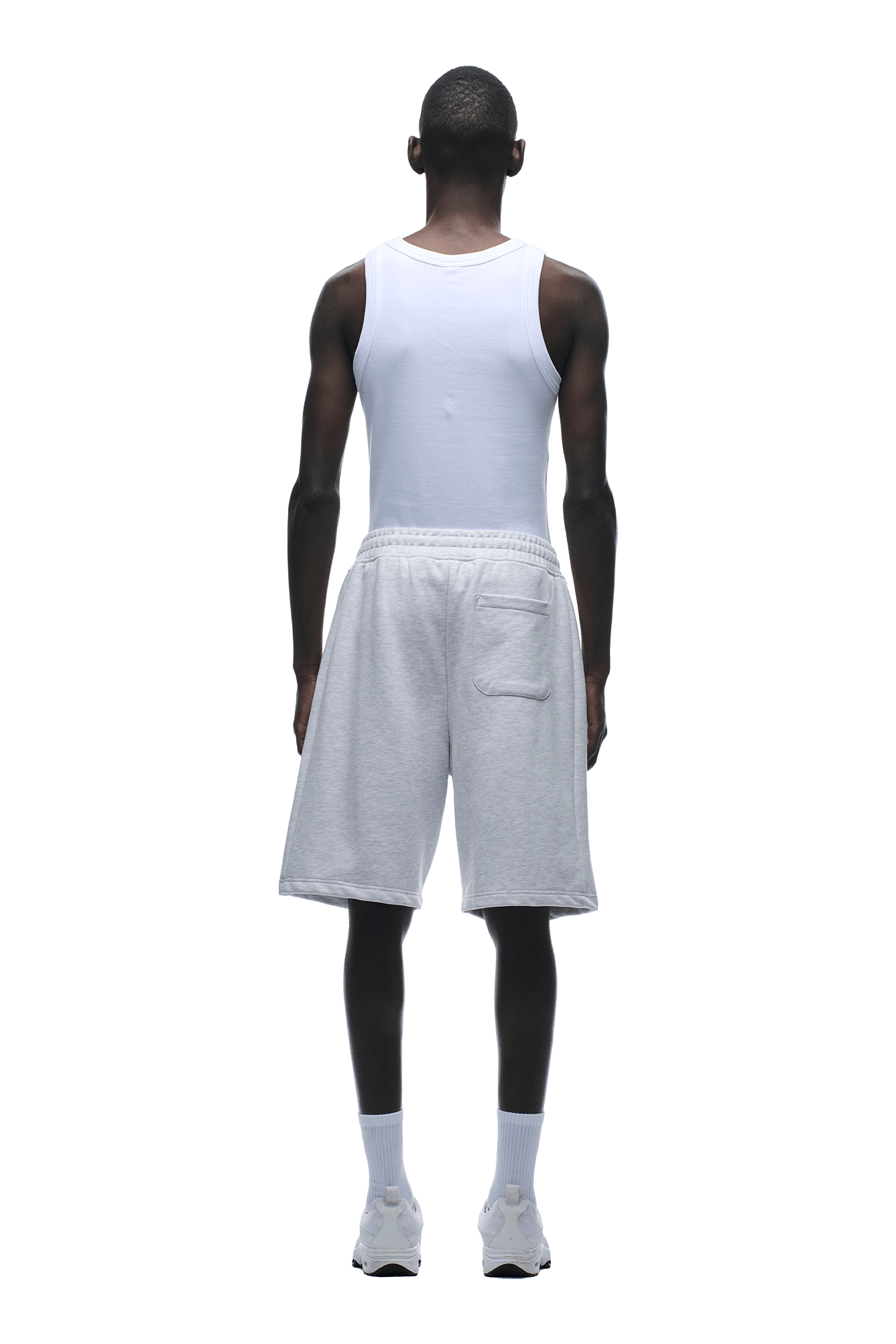 A man stands facing away against a plain green background, wearing a white tank top, 6PM 6PM+ SHORTS LIGHT GREY with a relaxed fit and drawstring waist, white socks, and white sneakers.