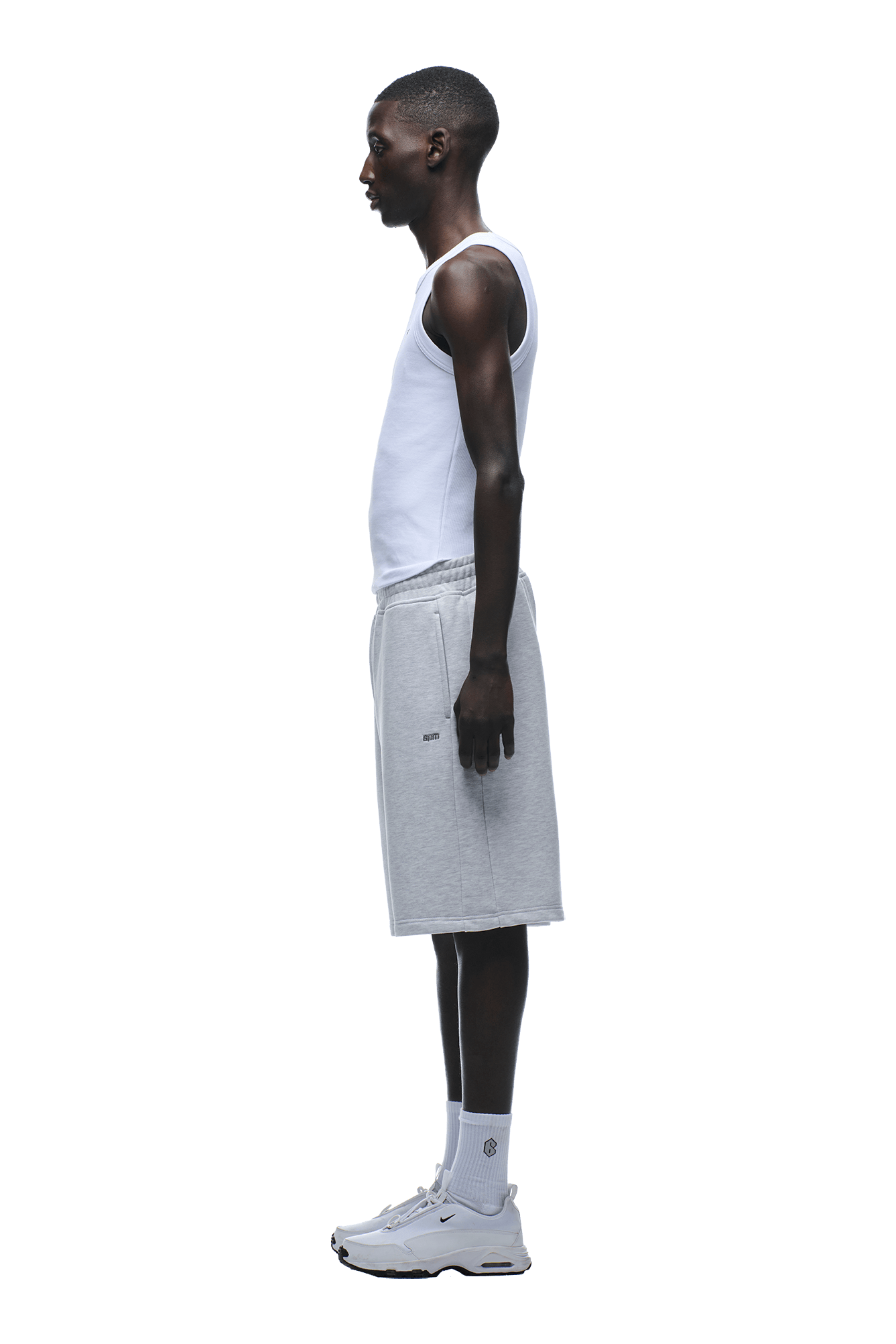 A man stands in profile against a plain green background, lounging in a white tank top, 6PM 6PM+ SHORTS LIGHT GREY with a drawstring waist, white socks, and white sneakers.