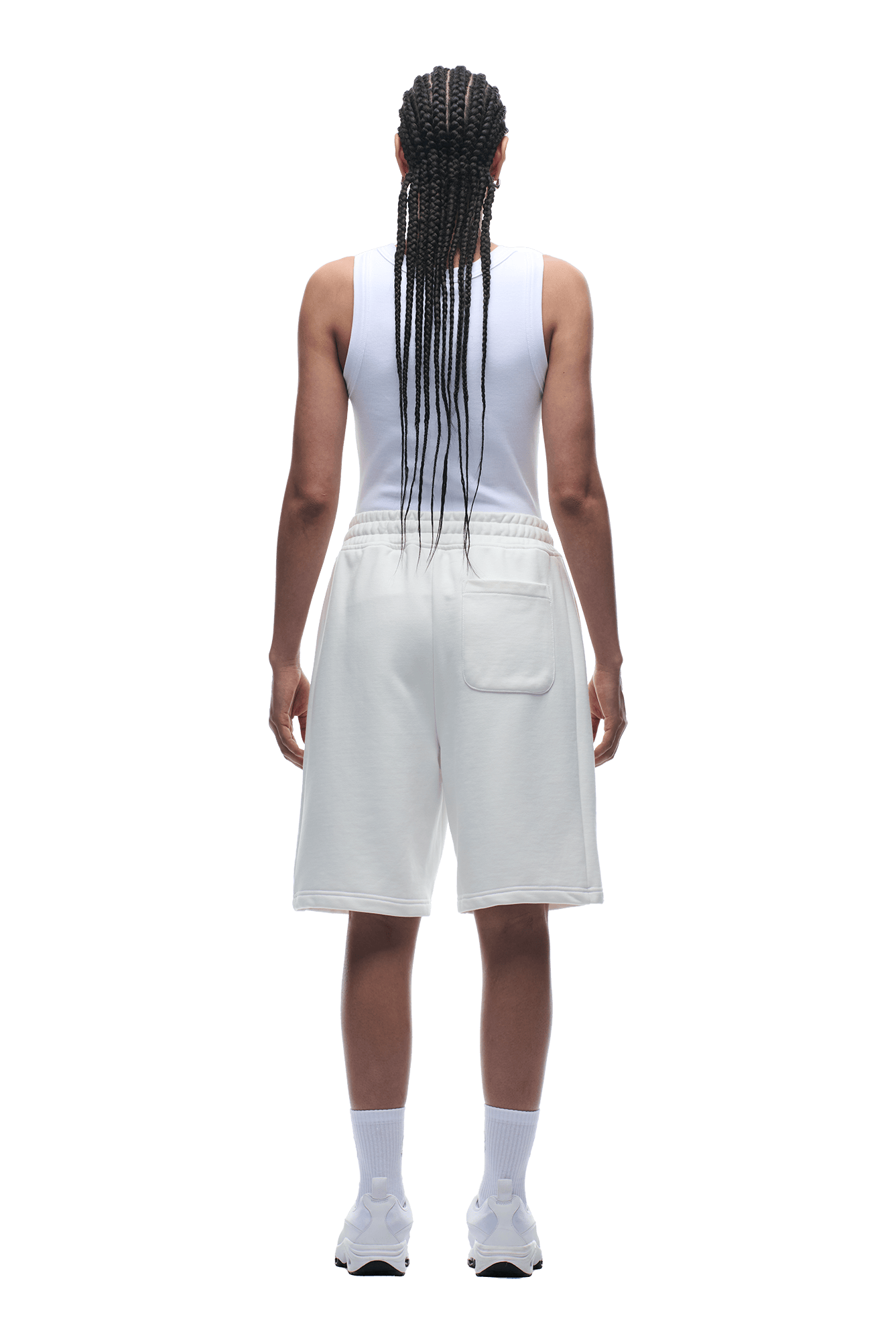 A person with long, braided hair stands facing away, wearing a white tank top and 6PM+ SHORTS COCONUT WHITE by 6PM, paired with white socks and sneakers against a transparent background.