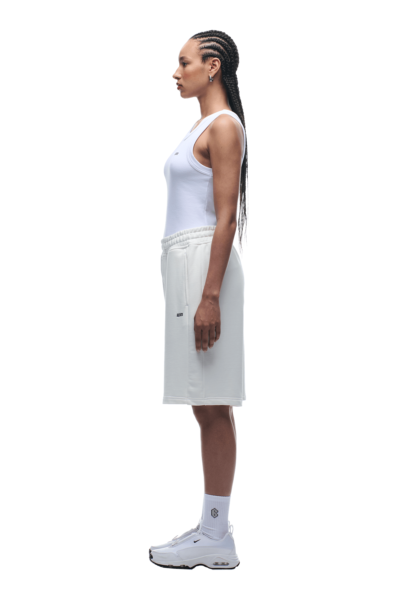 A woman with long braids, wearing a white tank top, 6PM+ SHORTS COCONUT WHITE by 6PM, white socks, and white sneakers stands in profile facing left against a plain green background.