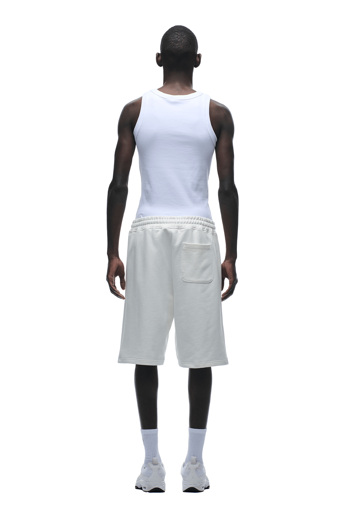 A person stands facing away in 6PM+ SHORTS COCONUT WHITE by 6PM, paired with a relaxed white tank, white crew socks, and sneakers, set against a plain green background.